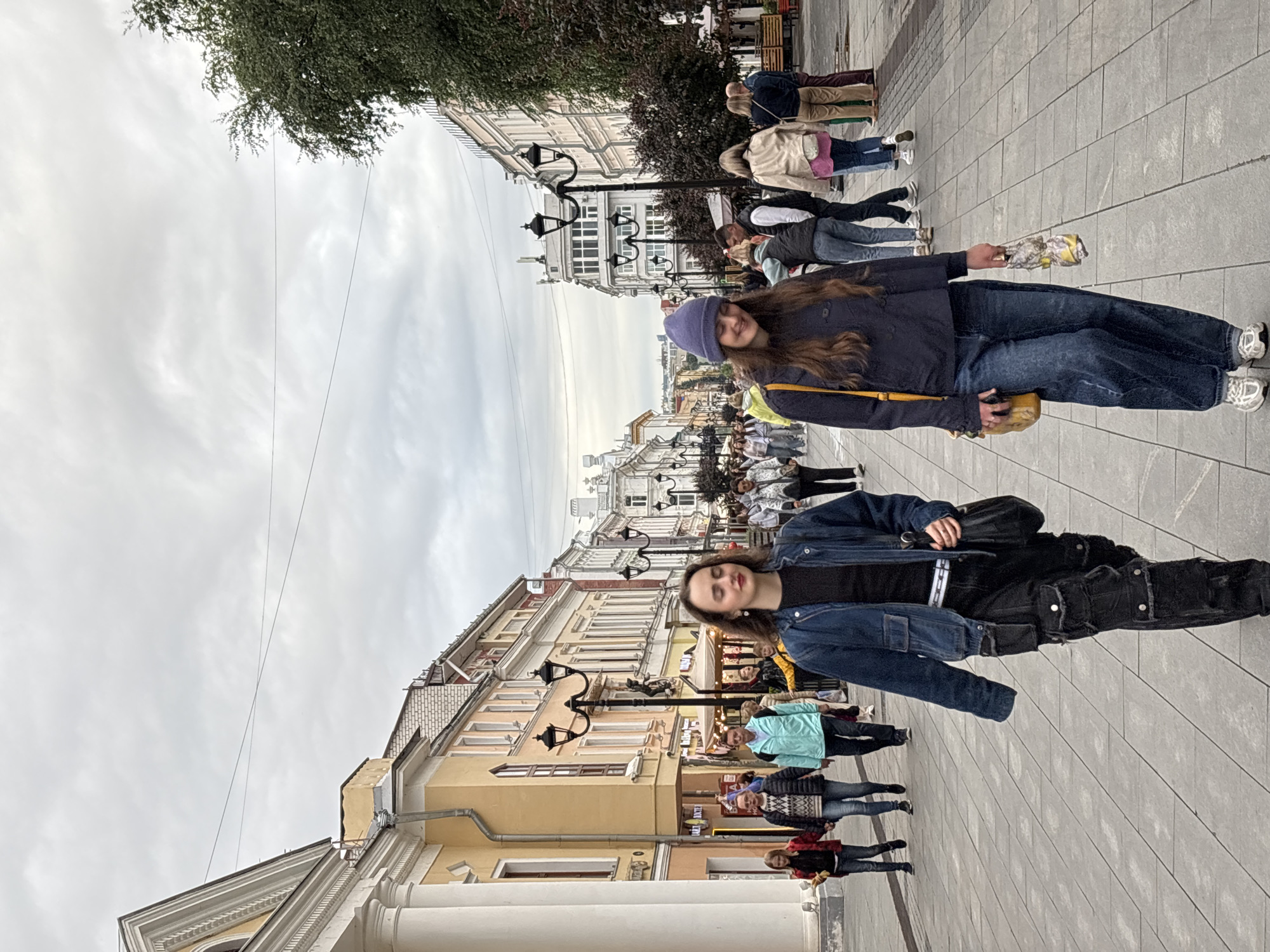 This is an eye-level, medium shot of two young women walking towards the camera on a pedestrian street in Nizhny Novgorod, Russia. The woman on the left is wearing a denim jacket, black shirt, and black cargo pants, carrying a black bag over her shoulder. The woman on the right is wearing a dark blue hooded sweatshirt, blue jeans, and a purple beanie, carrying a yellow bag and holding an umbrella. They are walking on a paved street lined with buildings on both sides. The buildings are a mix of architectural styles and colors, with some appearing to be shops or businesses. Other pedestrians are visible in the background, walking in both directions. The sky is overcast. The street appears to be Bolshaya Pokrovskaya Street, a well-known pedestrian street in Nizhny Novgorod.