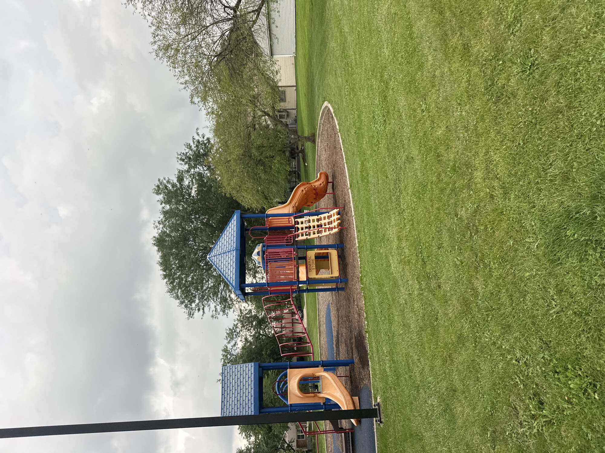 The image shows a playground located in Streamwood, Illinois, USA. The playground features a blue and red structure with slides, a climbing net, and a bridge connecting two platforms. The ground beneath the playground is covered in wood chips, and a grassy area surrounds it. In the background, there are trees and houses, suggesting a suburban setting. The sky is overcast. There are no people or animals visible in the image. The scene is static and appears to be taken during the daytime.