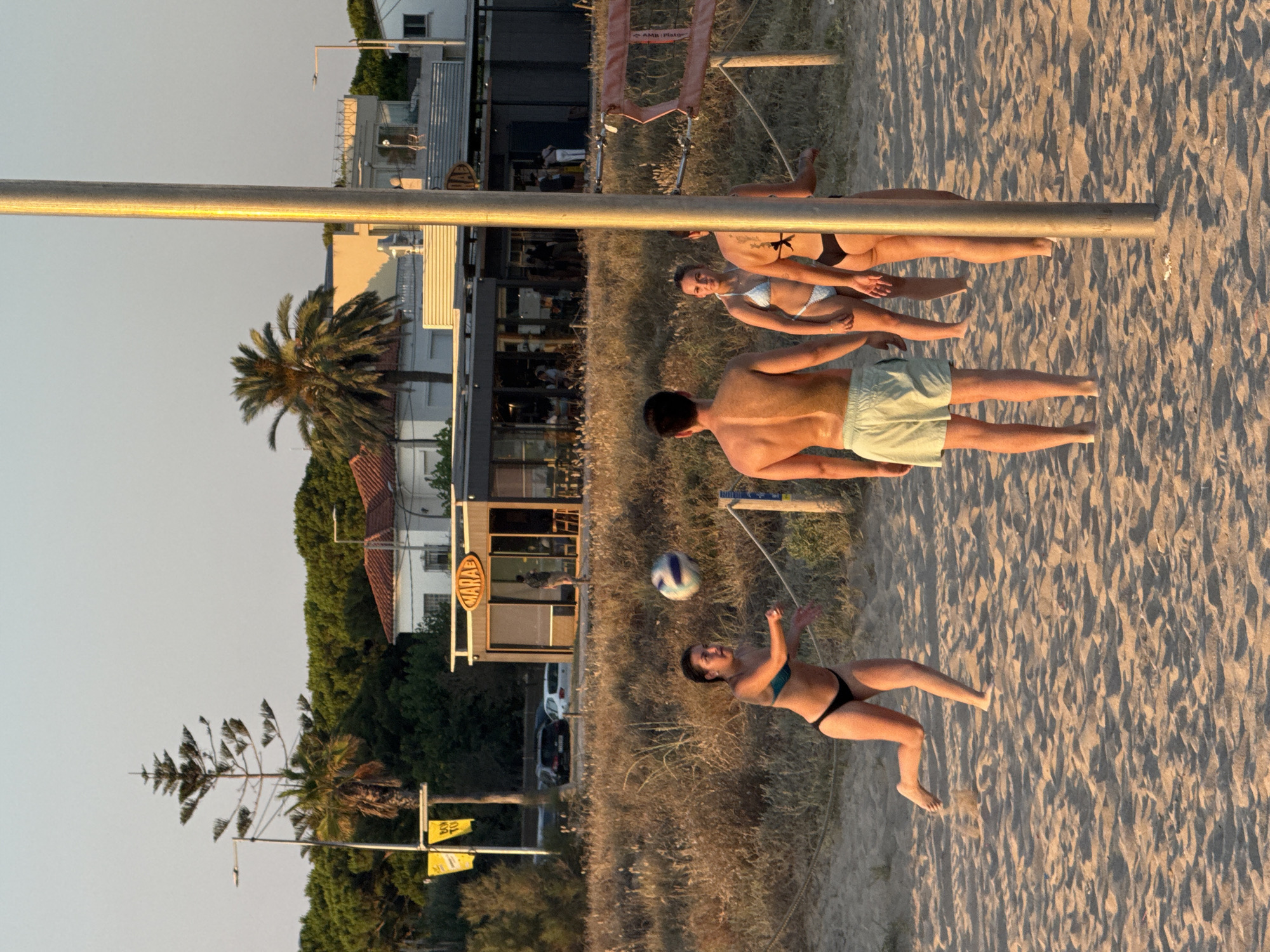 A group of young adults are playing beach volleyball on a sandy beach in Castelldefels, Spain. The scene takes place during the golden hour, with the warm sunlight casting long shadows across the sand, suggesting it is late afternoon or early evening. The sky is clear and pale, indicating fair weather.
In the foreground, a man wearing light green shorts is lying on the sand, his torso visible. Next to him, a woman in a black bikini is in the process of playing volleyball, reaching up to hit the ball. She appears focused and active. Further back, another woman, also in a bikini, is lying down on the sand, looking towards the sky. Behind her, two more individuals are partially visible, also relaxing or playing.
The background shows a building with a sign that reads "MARAE" in yellow lettering, suggesting a nearby establishment such as a restaurant or beach bar. Palm trees and other vegetation are visible behind the building, adding to the coastal atmosphere. A volleyball net is set up, confirming the activity. The overall mood seems relaxed and recreational, capturing a typical summer day at the beach.