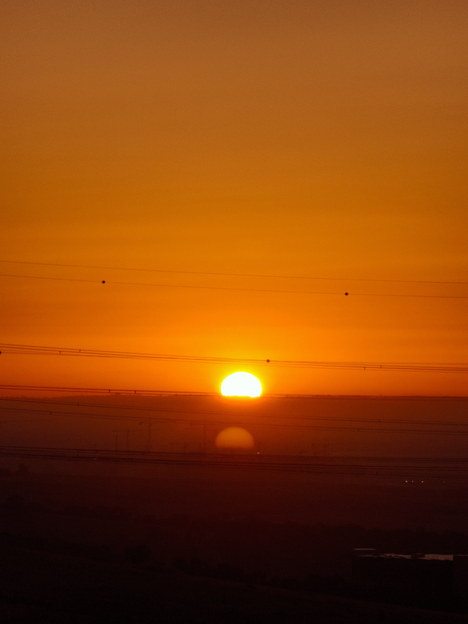 The image shows a sunset over Be'er-Sheva, Israel. The sun, a brilliant orange orb, is partially obscured by the horizon, casting a warm glow across the sky. Several thin, dark power lines stretch horizontally across the frame, silhouetted against the vibrant sky. In the lower portion of the image, the landscape is a dark silhouette, hinting at the city below. A faint reflection of the sun is visible on what appears to be a body of water in the distance. The overall impression is one of a peaceful end to the day, with the sun's descent creating a dramatic and colorful spectacle.