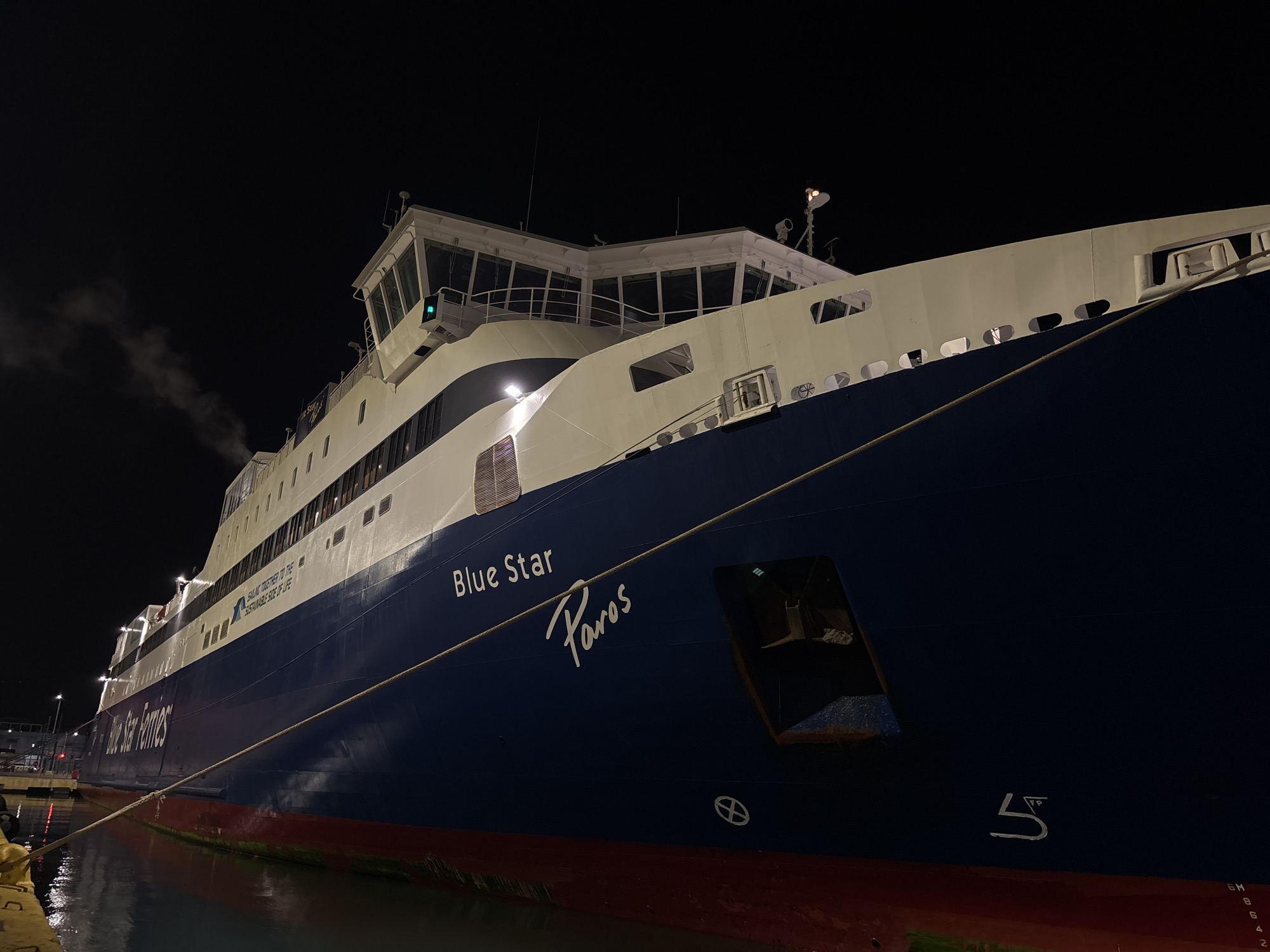 The image shows a nighttime view of the Blue Star Paros ferry docked at the Port of Piraeus, Greece. The ferry is a large, blue and white vessel with "Blue Star Paros" printed on its side. The bow of the ship is visible, showing the anchor housing and draft marks. A thick mooring rope extends from the ship to the dock. The background is dark, with some visible lights and structures from the port area. There is some smoke coming from the ship. The water in the foreground shows reflections of the ship and dockside lights.