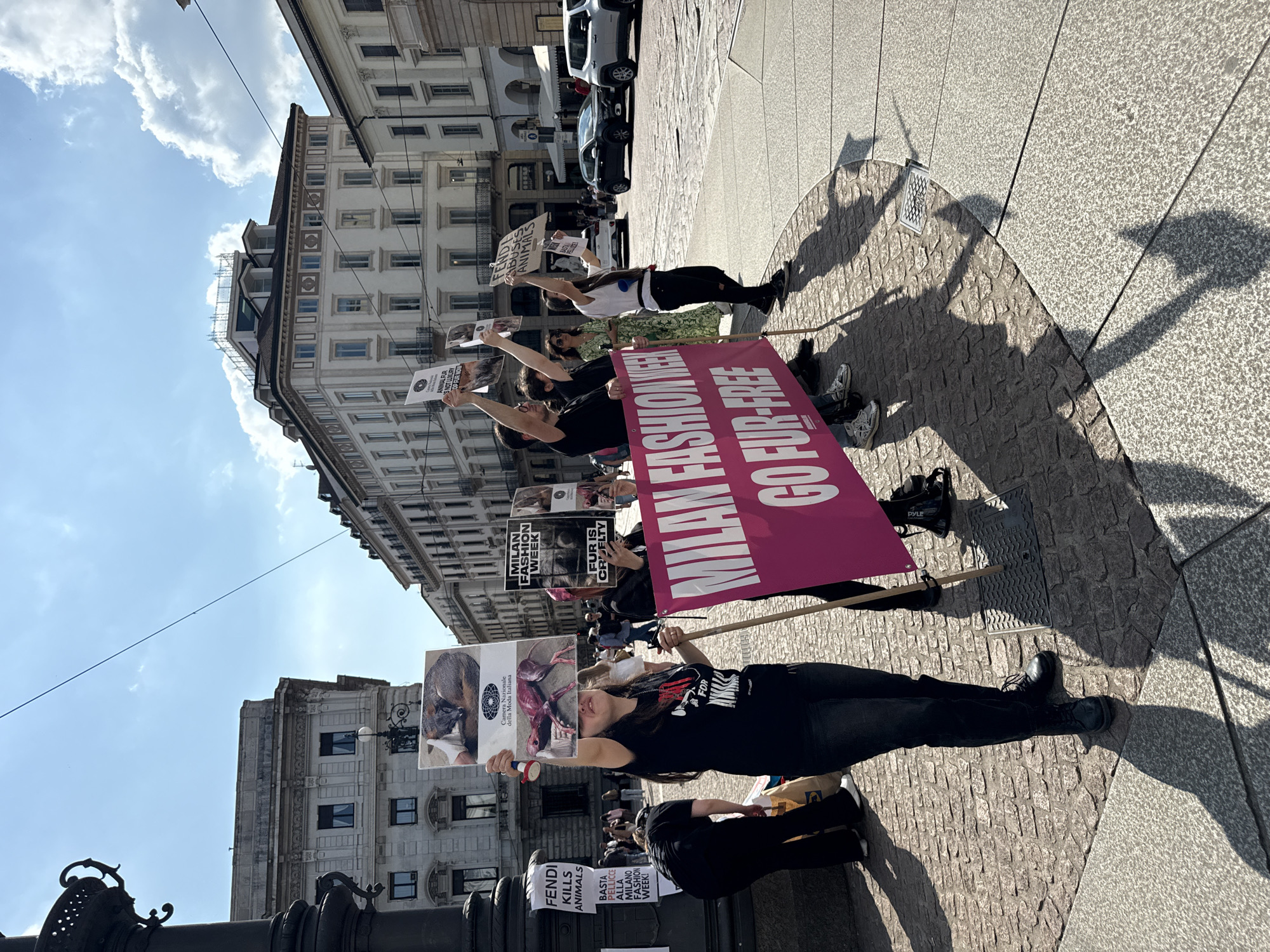 A daytime outdoor shot shows animal rights protestors in Milan, Italy, likely near a fashion venue, holding signs that critique Milan Fashion Week and companies like Fendi for the use of animal fur. One large pink banner reads "MILAN FASHION WEEK GO FUR-FREE." Other signs feature images of animals and messages such as "FUR IS CRUELTY," "FENDI KILLS ANIMALS," and "FENDI ABUSES ANIMALS." The protestors, dressed in casual clothes, hold signs aloft. The scene is set in a city square with buildings in the background. Shadows of the protestors are visible on the pavement. Several cars can be seen behind the protestors.