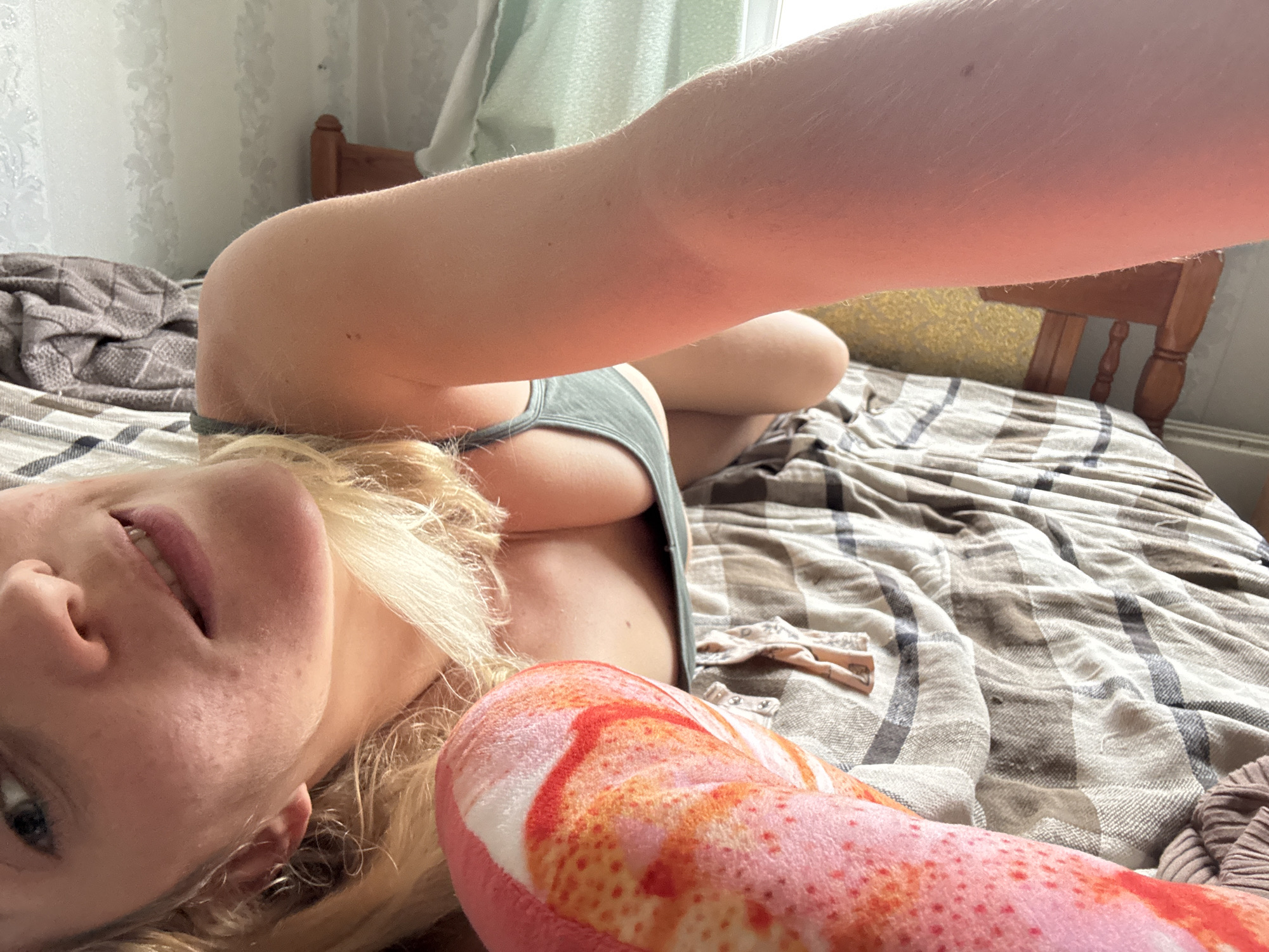 A bright, natural-light selfie captures a young woman lying on a bed in what appears to be a bedroom in Minsk, Belarus. The image is taken from an overhead perspective, likely by the woman herself, as her arm is extended holding the camera. She has blonde hair, fair skin with faint freckles, and a relaxed, smiling expression, showing a hint of her teeth. Her blue/grey eyes are looking towards the camera. She is wearing a light green or grey tank top or camisole.
In the foreground, a prominent pink and orange fluffy object, possibly a stuffed animal or a decorative pillow with a unique pattern, rests near her. The bed is covered with plaid bedding in shades of brown, grey, and white. Behind her, a wooden headboard or the back of a wooden chair is visible, along with a light-colored curtain or wall hanging, indicating an indoor setting during daytime hours, as suggested by the soft, ambient light. A small piece of light-colored clothing, possibly a baby's onesie, is also visible on the bed in the middle right of the frame. There is no visible text in the image.