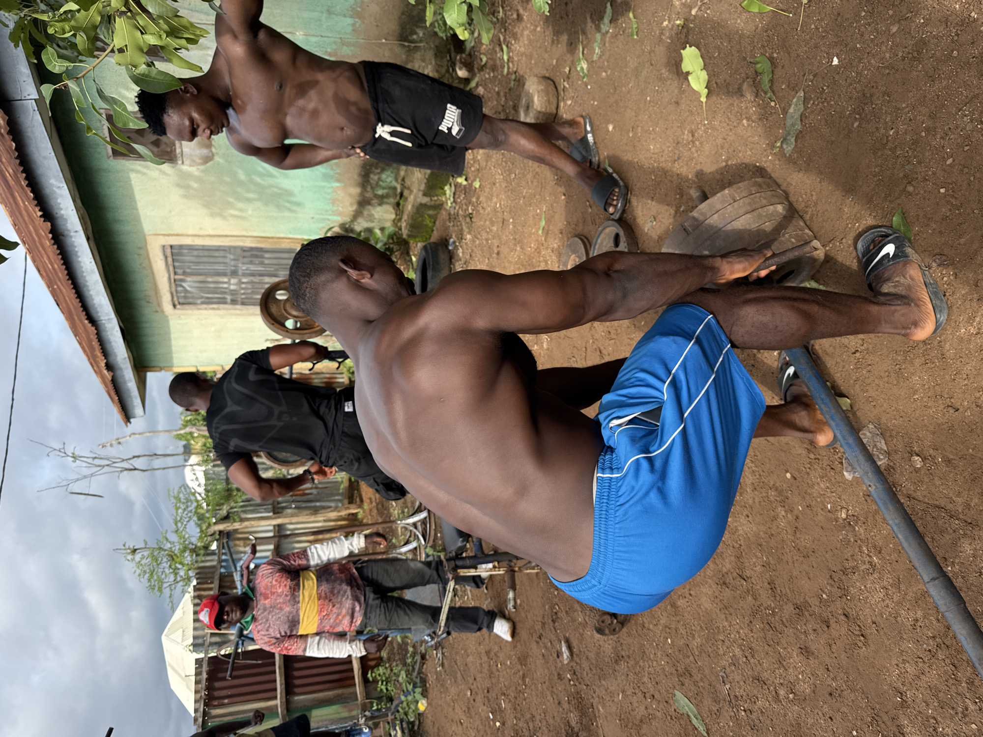 This image captures an outdoor, informal gym setting in Akwanga, Nigeria, under an overcast sky. The ground is composed of reddish-brown dirt, uneven and scattered with green leaves, dry plant debris, and a white piece of plastic waste. In the background, a single-story structure with faded and peeling light green/teal painted walls stands, topped by a rusted corrugated metal roof. It features two dark, rectangular window openings that appear to be covered or boarded up, and a low concrete foundation. To the left, a section of corrugated metal fencing (brown and green) is visible, with a white tent or roof structure partially seen beyond it. Sparse vegetation, including a prominent leafy branch, frames the scene.
Four adult males are present, engaged in or observing physical exercise. In the foreground, a muscular, dark-skinned male with short-cropped hair wears blue athletic shorts with white side stripes and black Nike slide sandals. He is bent forward, with his back to the viewer, actively gripping a dark metal bar with his right hand, in the process of lifting or lowering a large, circular weight plate that rests on the ground. Several similar dark, circular weight plates are scattered on the dirt near his feet.
To the right, another muscular, dark-skinned male with short dark hair stands with his hands on his hips, facing towards the left foreground. He wears black athletic shorts featuring a white drawstring and a white "PUMA" logo (with a book graphic) on the left thigh, along with black Nike slide sandals. A flat, round stone or weight rests on the ground beside his right foot.
Behind the foreground figures, closer to the green building, a third dark-skinned male is partially visible. He wears a black short-sleeved t-shirt with a subtle textured pattern and black shorts. He is facing away from the viewer and appears to be interacting with a large circular weight plate and a segment of metal exercise equipment, possibly a weight rack.
Further to the left, a fourth dark-skinned male wears a red baseball cap, a long-sleeved t-shirt with a multi-colored pattern (brown, red, yellow, white), dark trousers, and white athletic shoes. He stands upright, looking towards the viewer, appearing to observe the activities. A piece of metal exercise equipment, possibly a bench, is situated behind him.
The scene depicts a raw, functional environment dedicated to strength training, utilizing basic and assembled weightlifting equipment.