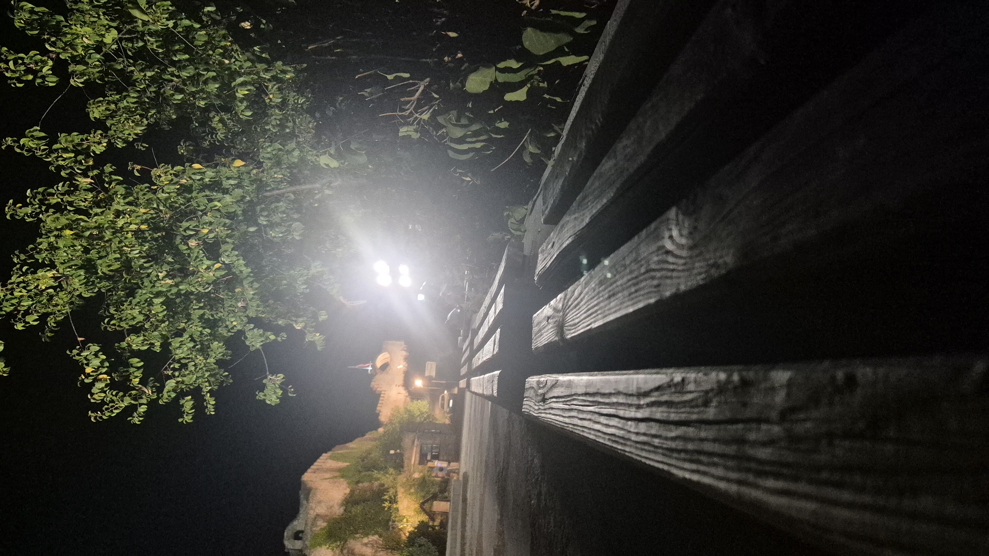 The image captures a nighttime scene at the Lovech Fortress in Lovech, Bulgaria. In the foreground, a wooden bench is visible, its weathered planks illuminated by artificial lighting. The bench is oriented diagonally, leading the viewer's eye towards the background. Beyond the bench, a pathway or road extends into the distance, where the Lovech Fortress is prominently featured. The fortress, constructed from stone, is lit by several spotlights, casting a warm glow on its ancient walls and battlements. A flag, possibly the Bulgarian flag, is visible atop one of the fortress structures. In the mid-ground, a car is parked near the entrance to the fortress, and a few figures, presumably visitors, are visible near the entrance. Lush green foliage frames the scene on the right side, with a large tree whose leaves are illuminated by the bright lights. The overall ambiance is one of historical grandeur set against the darkness of night, enhanced by the strategic lighting of the fortress.