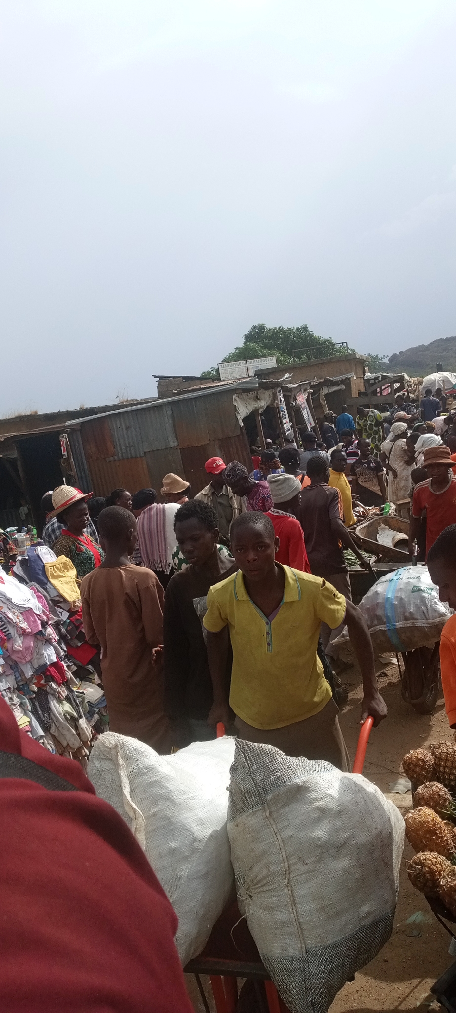 The image depicts a crowded marketplace in Gura Top, Nigeria. The scene is bustling with activity, showcasing numerous individuals engaged in commerce. People are densely packed, navigating through makeshift stalls and displays of goods. Several individuals are pushing wheelbarrows laden with merchandise, including large sacks and pineapples. Clothing is piled high in one area, suggesting a garment market. The buildings appear to be simple structures, some with corrugated metal roofs. The overall atmosphere is one of a vibrant, lively open-air market.
