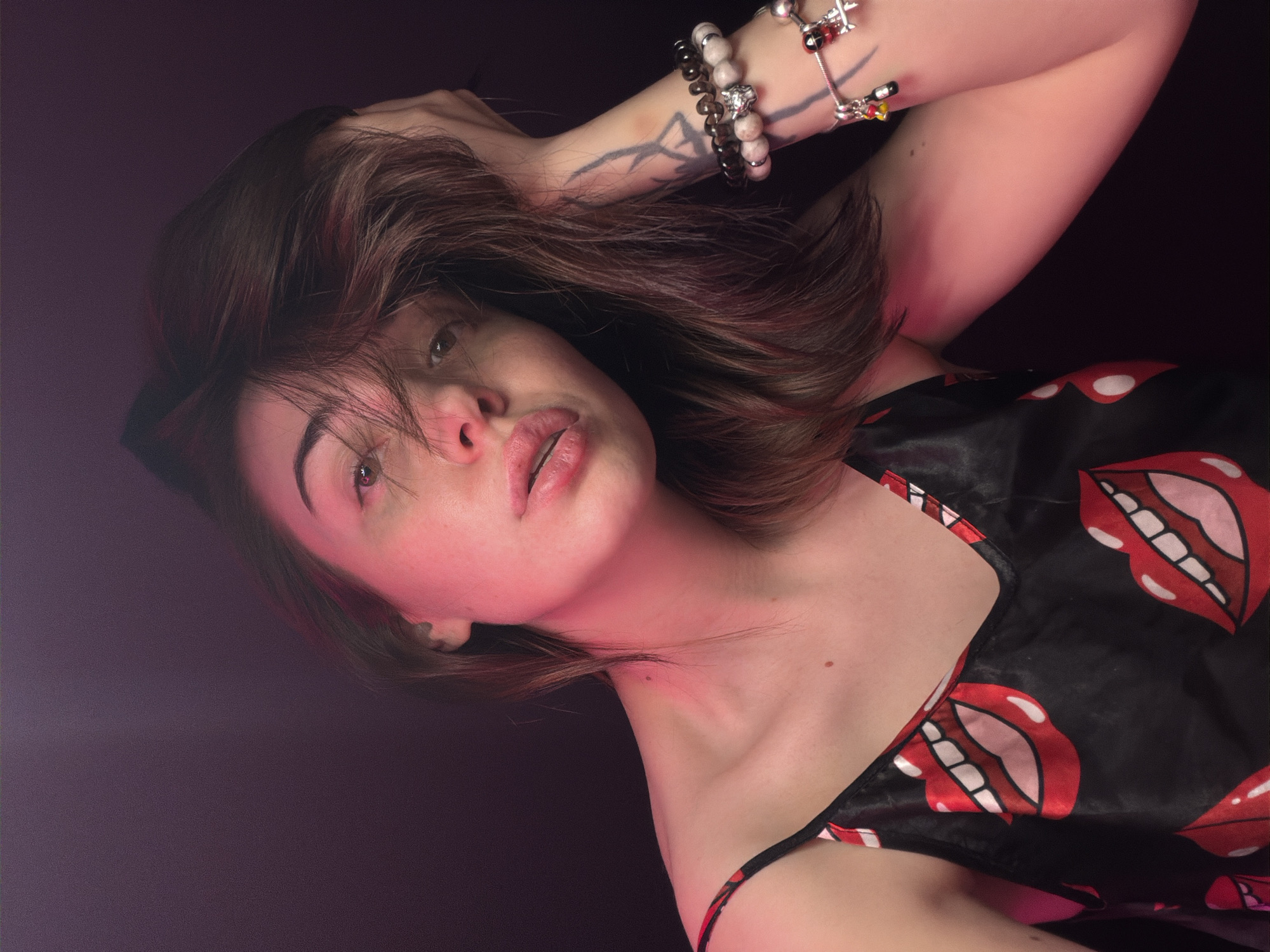 A young woman is photographed from the chest up, leaning her head on her left hand and resting her arm on her head. She is wearing a black spaghetti strap camisole with a red and white lip pattern. Her hair is dark brown and falls across her face, obscuring part of her left eye. Her right eye is visible, and she has fair skin with light makeup. A tattoo is visible on her left forearm, along with several bracelets. The background is dark and out of focus, with a pinkish-purple light casting a glow on the left side of her face and neck.