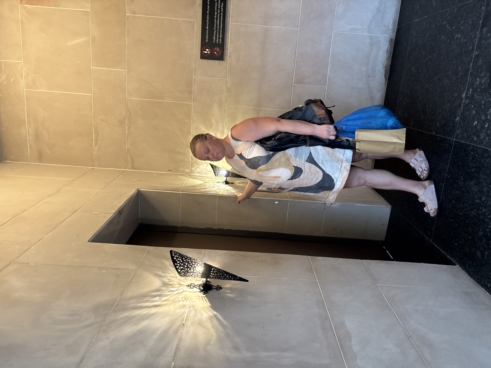 A light-skinned woman with blonde hair pulled back stands in front of a wall in what appears to be a modern building, possibly a hotel or elevator lobby. The wall is made of large, light-colored stone tiles, and there is a dark opening to her left, suggesting an elevator or doorway. The woman wears a sleeveless, light-colored dress with a geometric pattern in black, white, and beige. She is carrying a black bag with brown leather accents on her right shoulder and a brown paper bag and a blue plastic bag in her left hand. She is wearing light pink slip-on sandals. To the right of the woman, on the wall, is a black sign with white text and symbols, indicating rules and regulations. One of the signs states "Alcohol not allowed below 18 years of age" and information about drug trafficking. The lighting in the scene comes from decorative wall sconces on the left, casting interesting shadow patterns on the wall. The floor is dark and appears to be tiled.