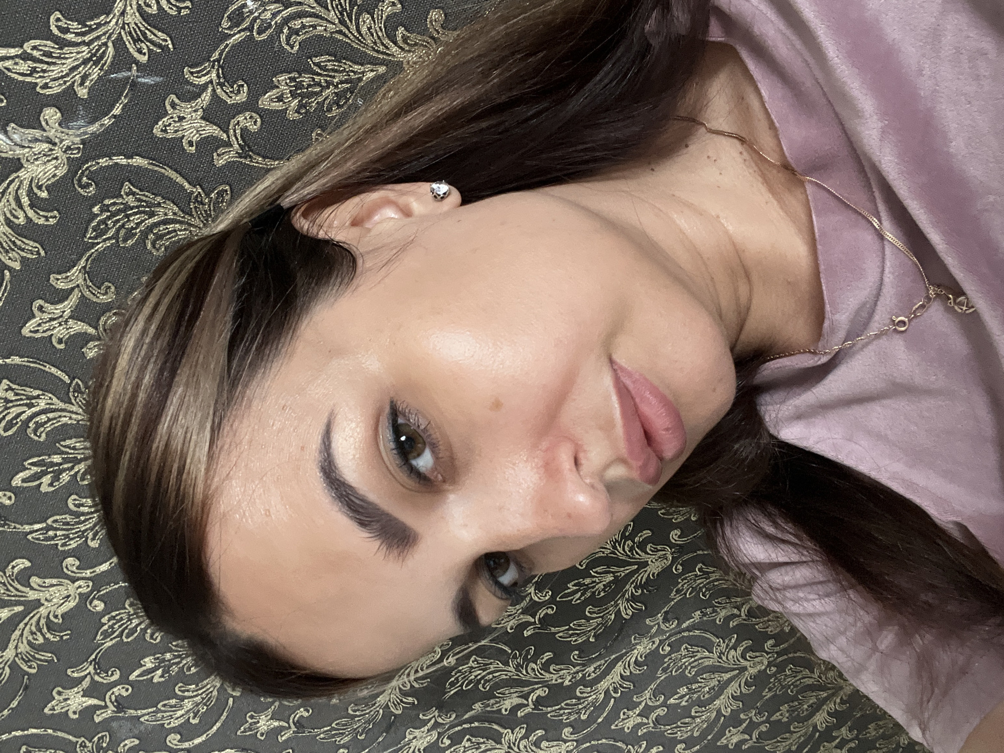 A medium close-up shot focuses on the face and upper chest of a woman with brown hair and light skin. She is looking towards the camera with her head tilted slightly. Her makeup includes defined eyebrows, subtle eye shadow, and a light pink lipstick. She is wearing a light pink, possibly velvet, top and a delicate gold necklace with a small pendant. A small stud earring is visible on her right ear. The background is a dark gray wallpaper with a repeating ornate pattern in a gold-like color. The lighting appears to be soft, highlighting the woman's features and creating a subtle sheen on her skin.