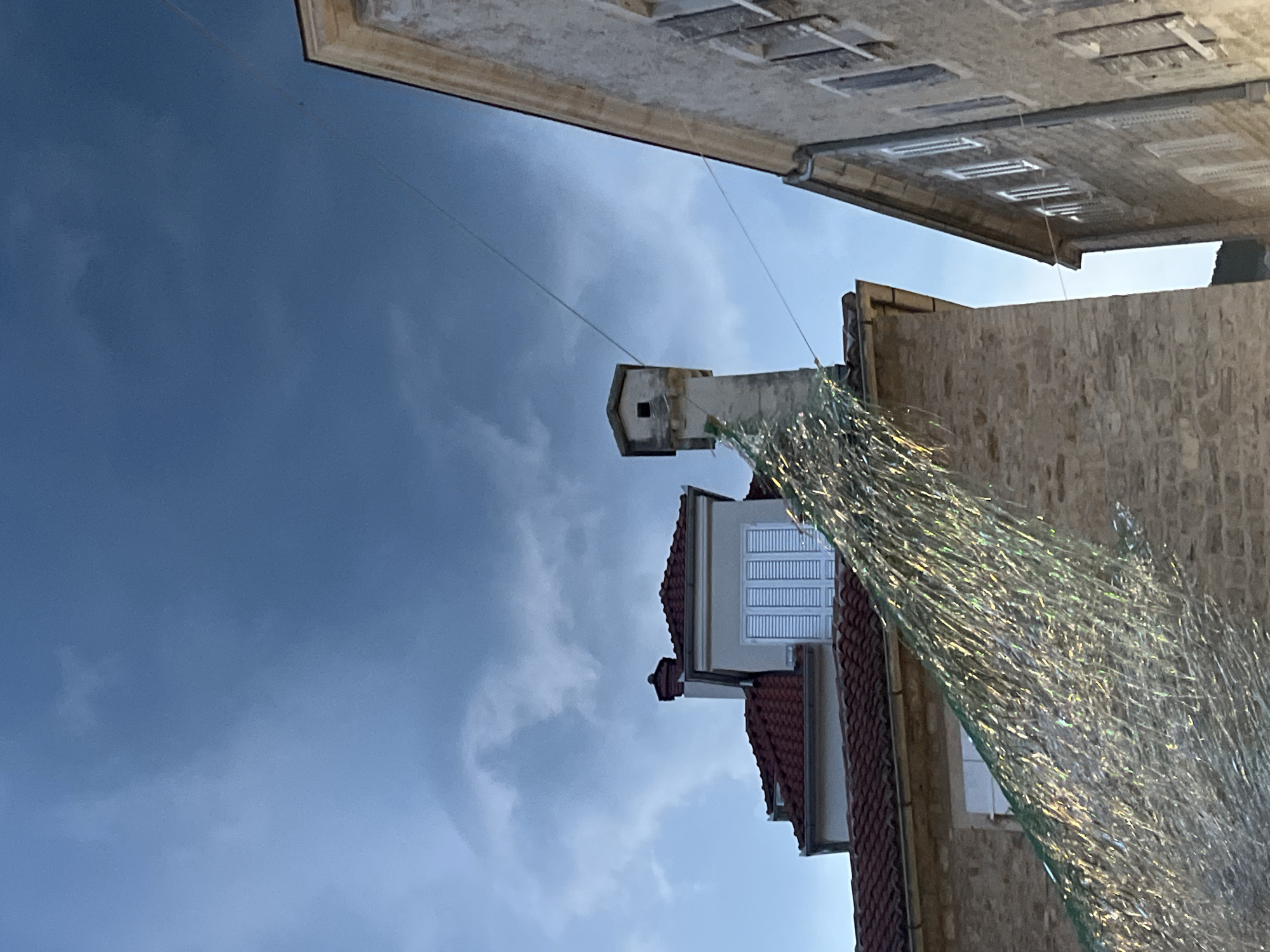 The image shows a close-up, upward-facing view of buildings against a dramatic sky. The perspective is disorienting, with the buildings appearing to extend upwards from the bottom of the frame. In the foreground, a shimmering, metallic netting or tinsel-like material is draped across the right side of the image, catching the light and creating a glittery effect.
Behind the netting, a textured brick wall of a building is visible. Above this, a section of a lighter-colored building with a red tiled roof and white-shuttered windows is seen. Further up, a narrow, stone structure with a small square opening, possibly a chimney or ventilation shaft, stands against a dark, cloudy sky. A thin wire stretches from this structure towards another building at the top of the frame. This top building features a pale facade with rows of recessed rectangular openings, suggesting windows or architectural details.
The sky is a mix of dark, brooding clouds and lighter, brighter areas, hinting at an impending storm or the aftermath of one. The lighting suggests it is either early morning or late evening, with the sun perhaps breaking through or just set. The overall impression is one of an urban setting, likely in Budva, Montenegro, given the architectural style and the presence of Mediterranean-style rooftops and stonework. No people are visible, and the focus is on the architectural elements and the atmospheric sky. There is no visible text in the image.