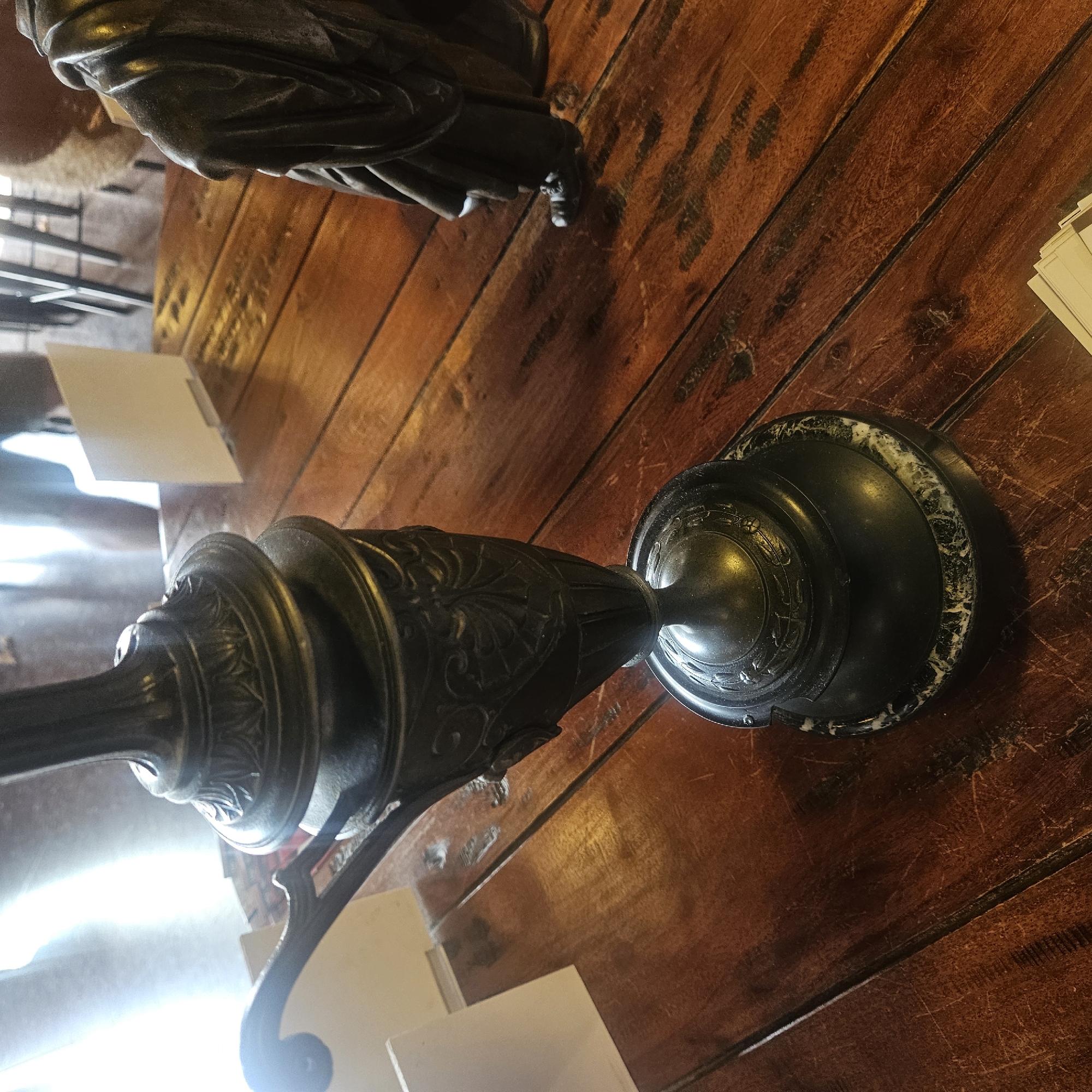 The image depicts a close-up, eye-level shot of a dark, ornate metal lamp base with a marble accent on a polished wooden table. The lamp has intricate carvings and a scrolled handle. To the right of the lamp base, a dark, cloaked figure, likely a statue or a mannequin, is visible from the knees down, standing on the same wooden surface. The background is blurred, showing a brightly lit area with what appear to be chairs and possibly other tables, suggesting an indoor setting such as a restaurant or cafe. There is a small white card or sign standing upright on the table in the middle ground. The overall scene is indoors and appears to be a quiet, possibly historical setting. The specific location of Almadrones, Spain, is not discernible from the visual information provided in the image itself.