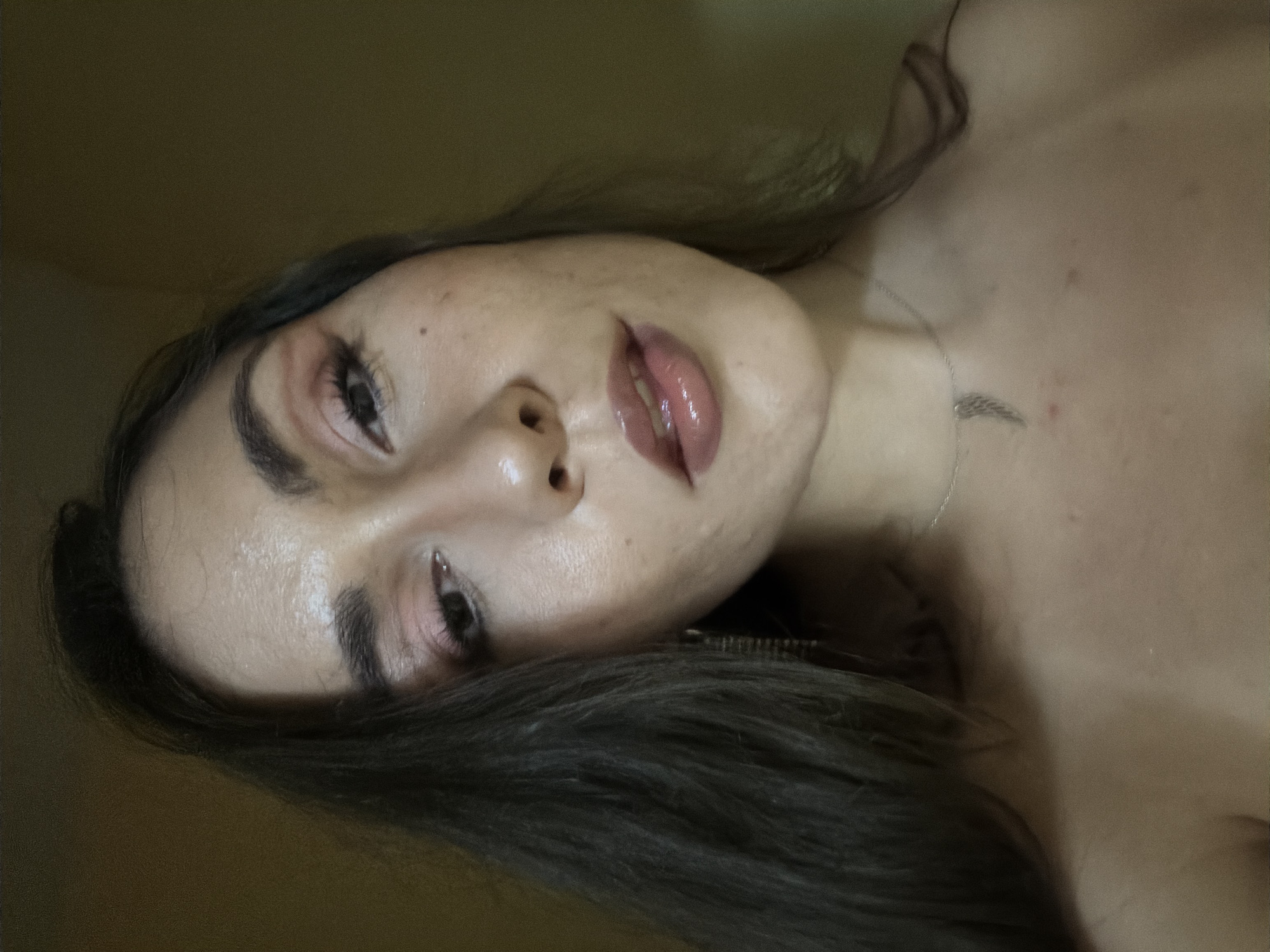 A close-up, head-and-shoulders shot of a young woman with dark hair, fair skin, and light brown eyes. She is wearing makeup, including eyeshadow and lip gloss. She has a delicate silver necklace with a wing pendant. The background is a plain, yellowish-brown wall. The image is taken indoors, likely in a residential setting, and there are no contextual clues to suggest a specific location in Varna, Bulgaria.