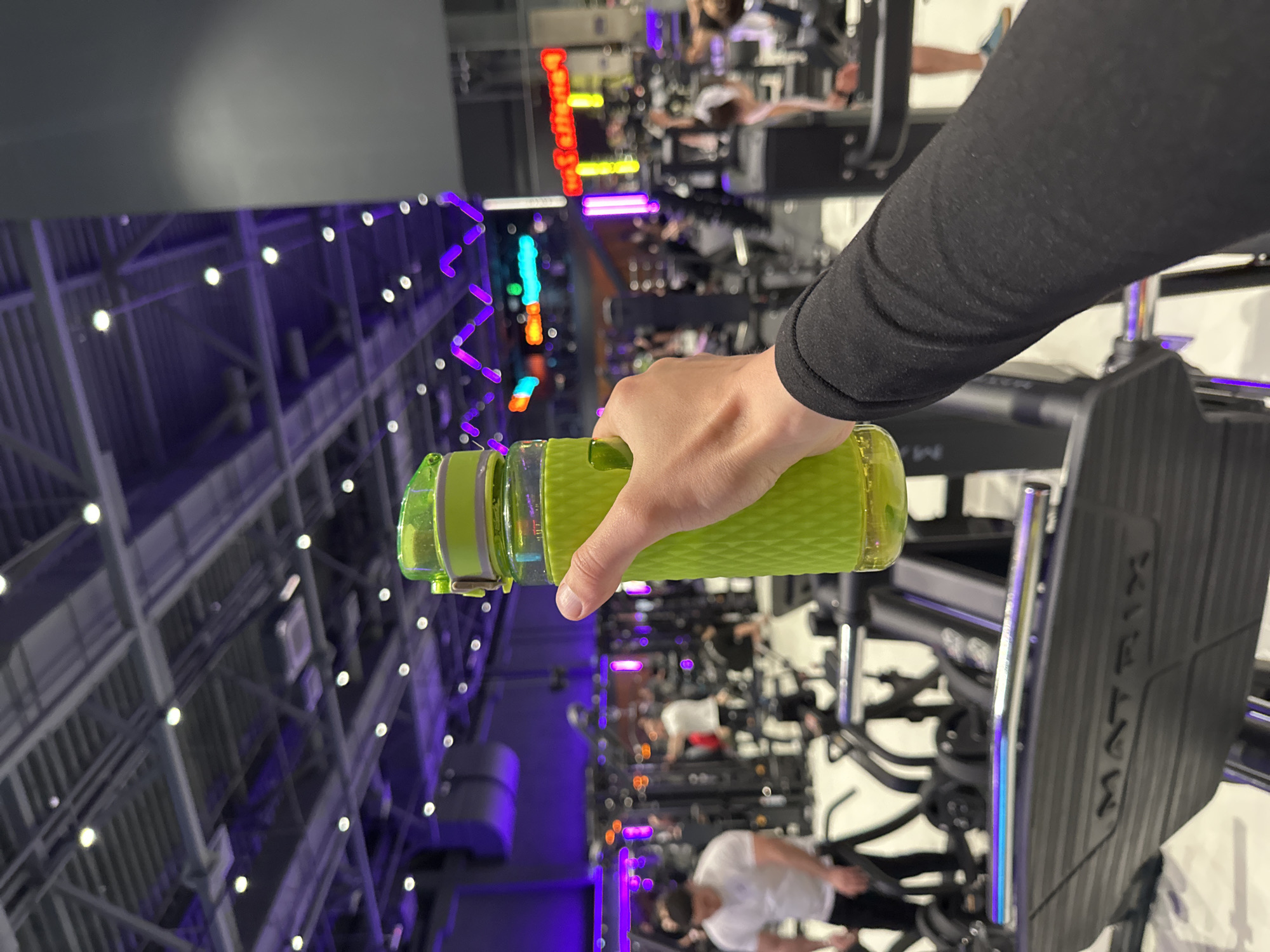 A hand holds a green water bottle inside what appears to be a brightly lit gym in Murino, Russia. The gym equipment, including a Matrix machine, is visible in the background, along with other people exercising. The gym features distinctive lighting, including neon signs and purple/blue lighting fixtures.