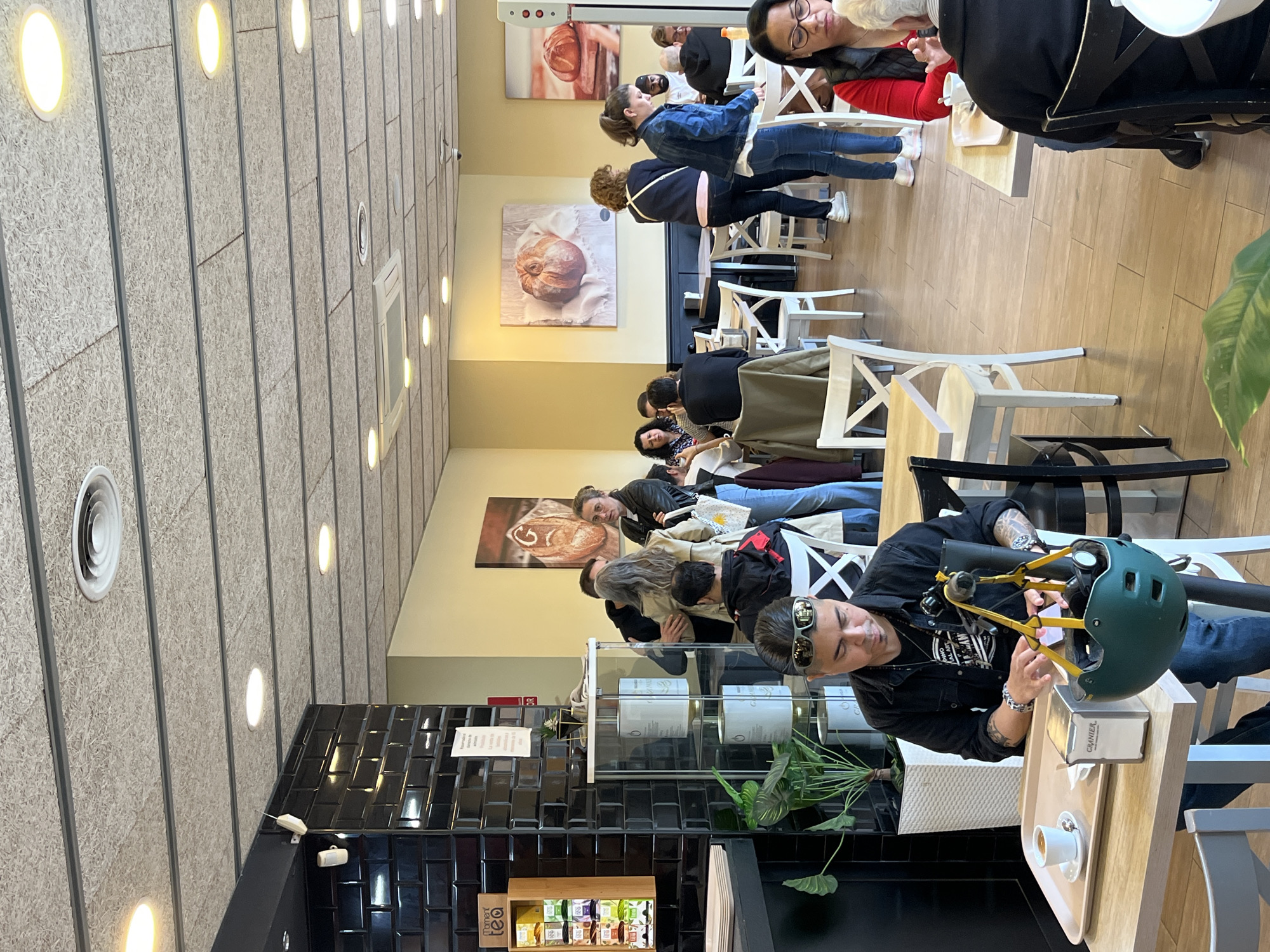 Here's a description of the image:
**Scene:** Interior of a cafe.
**People:** There are multiple individuals present. A person wearing a black shirt and sunglasses is sitting at a table and appears to be adjusting the straps of a helmet. Several other people are seated at tables in the background.
**Objects:** The foreground features a wooden table with a tray containing a coffee cup and a sugar packet. A green helmet is on the table. Other tables and chairs are visible in the background, along with paintings of bread on the wall. There is a display case with teabags, and plants are near the case and table.
**Environment:** The cafe has a modern design. The walls have tiled sections and textured panels with integrated lighting. Based on the architectural style and the types of food/drinks advertised, it could be located in Barcelona, Spain.