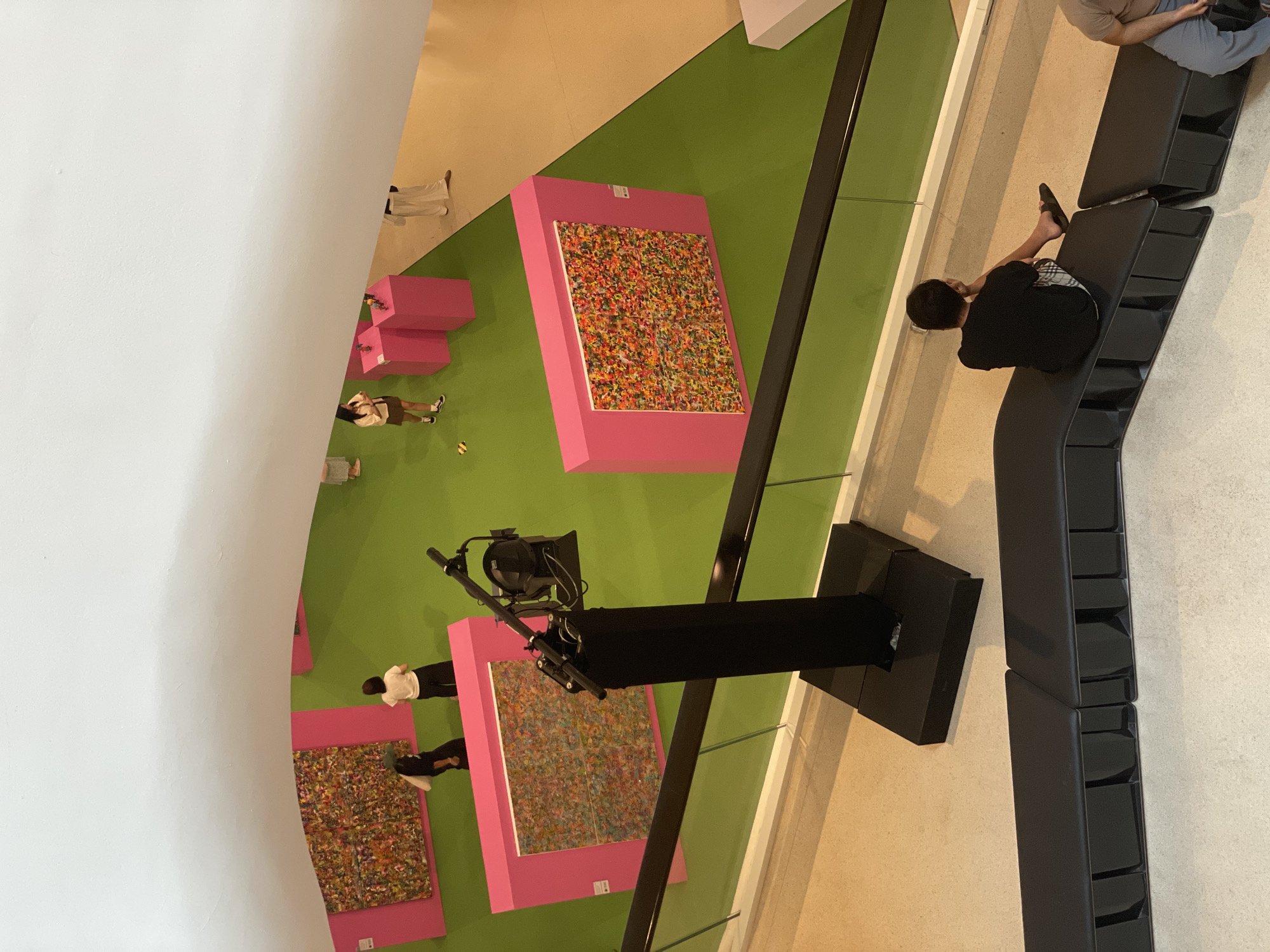 The image provides a high-angle, top-down view of an art exhibition space, likely an indoor gallery in Bangkok, Thailand. The floor is distinctly divided into a large, vibrant green area in the center and a lighter beige/tan section on the right, contrasted by a sweeping, curved white wall on the left.
Several visitors are present. On the beige floor to the far right, a person wearing a black t-shirt and plaid shorts is seated casually on a long, dark grey, segmented bench, with their bare feet visible. Another individual is partially visible next to them on the bench. On the green floor, people are scattered, observing the exhibits or moving around. One person in a light top and dark skirt/shorts stands near the white wall, while another, wearing white attire, is visible closer to the beige floor. Further down, a person in a white top and black pants stands near one of the artworks.
The main objects of interest are the artworks themselves: large, rectangular pieces displayed on bright pink pedestals or stands. The artworks are abstract and highly colorful, appearing to be composed of numerous small, multi-hued elements resembling confetti or sprinkles. A prominent black structure, possibly a camera or lighting rig with an attached spotlight or projector, rises from the green floor near the center-right of the frame. A small yellow object is visible on the green floor near one of the observers.
The activity suggests a casual visit to an art exhibition, with people viewing the displays and resting. The scene is brightly lit by what appears to be artificial indoor lighting, indicating an indeterminate time of day. No explicit text is clearly visible on any of the artworks, pedestals, or surrounding elements.