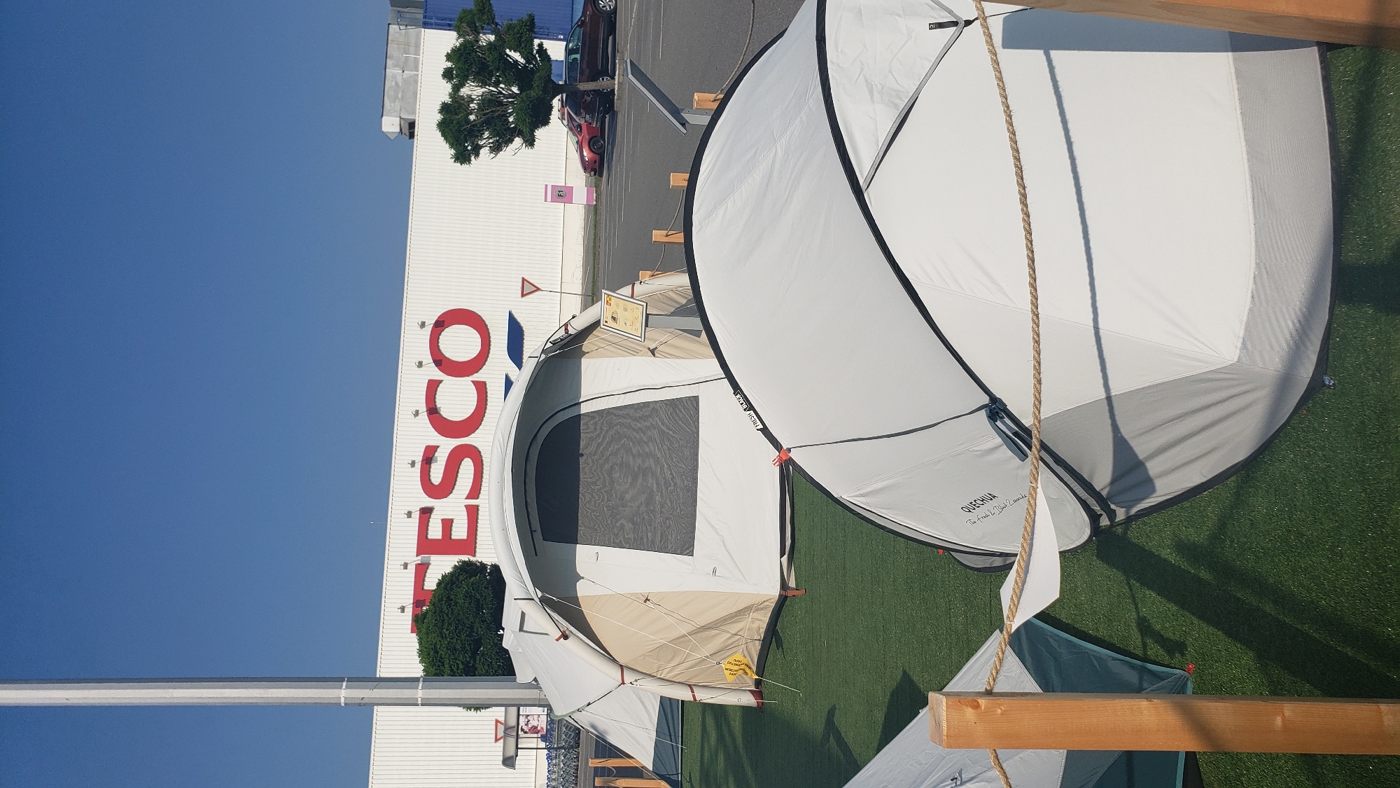 This image depicts a sunny outdoor scene in Prešov, Slovakia, featuring a large white building with red "TESCO" lettering. In front of the building, several tents are set up on a green artificial turf surface. A tall, grey pole stands to the left of the frame. In the middle ground, a beige and white tent with a dark mesh window is visible, alongside a small display sign. A white pop-up tent with "QUECHUA" branding is prominently featured in the foreground. In the background, a parking lot is visible with a red car and a dark car. A small, decorative tree stands to the right of the Tesco building. The sky is clear and blue.