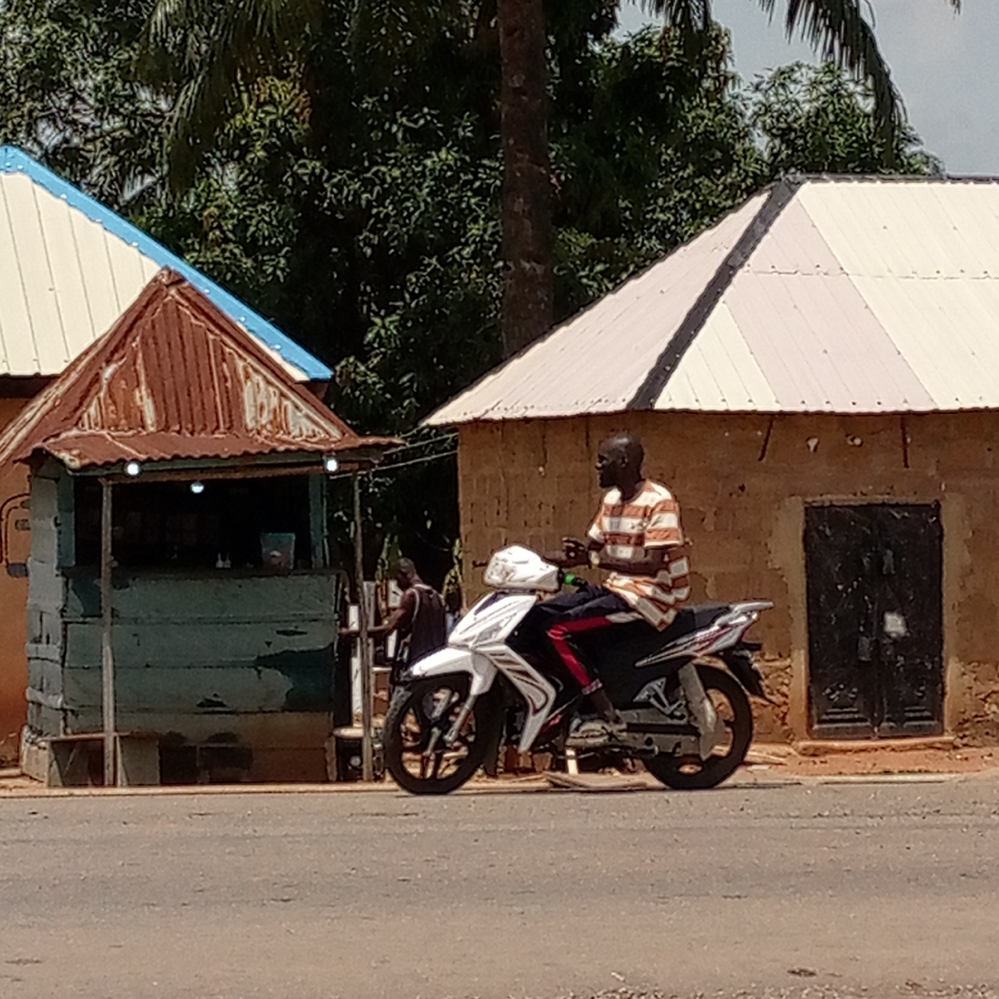 A man in a striped shirt sits on a white motorcycle on a road in Mararaba, Nigeria. Behind him, a person stands at a small, weathered green building with a rusty roof. Adjacent to the small building is a larger building with a white roof and a dark-colored door. Trees and other foliage are visible in the background.