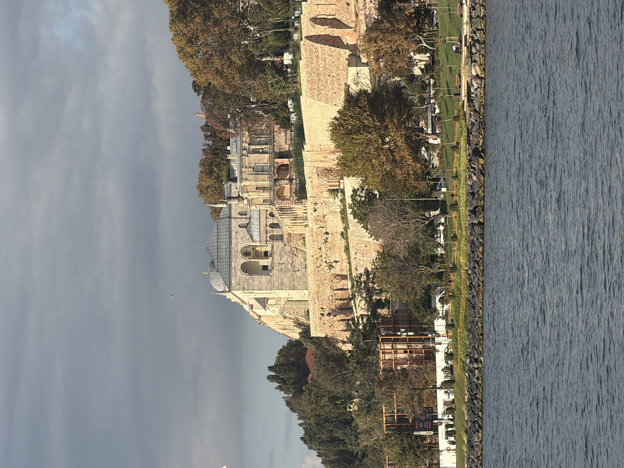 The image depicts a view of the Topkapi Palace complex in Istanbul, Turkey, from across a body of water, likely the Bosphorus strait. The palace complex, characterized by its stone architecture and domed roofs, is situated on a hill overlooking the water. The structures are partially surrounded by lush greenery and trees, some of which show autumn foliage. The foreground features the rippling surface of the water, bordered by a rocky embankment. A few scattered birds are visible in the sky. The scene is captured under a partly cloudy sky, with sunlight illuminating parts of the palace and its surroundings. No people are clearly discernible in the image.