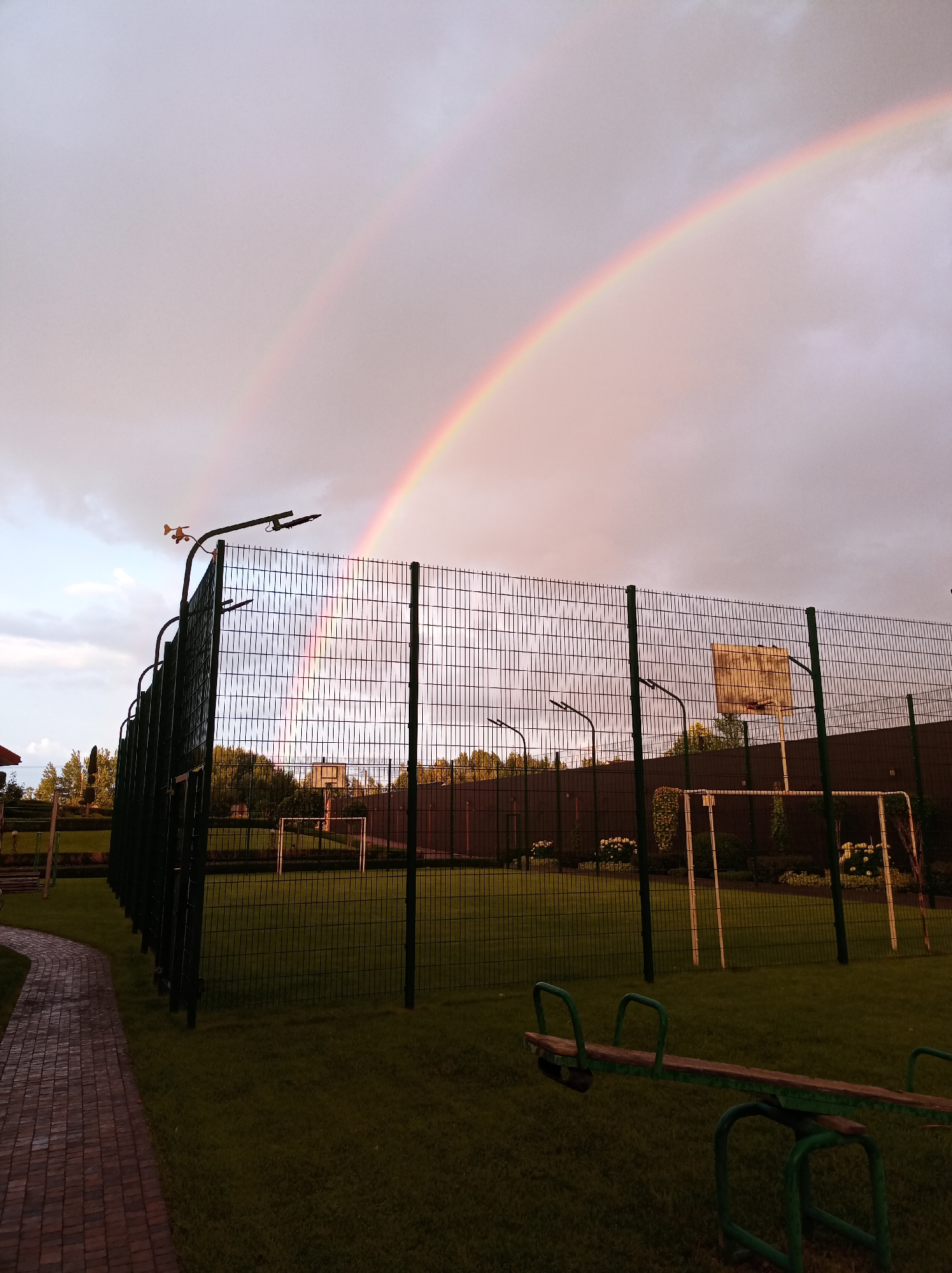 The image depicts a backyard area in Kozyn, Ukraine, with a double rainbow arching across a cloudy sky. In the foreground, a green seesaw is positioned on a lush green lawn. A paved walkway curves into the frame from the left. A tall, dark green metal fence encloses a sports field, where a basketball hoop and a soccer goal are visible. Trees and other structures are visible beyond the fence. The overall scene suggests a post-rain atmosphere, illuminated by the soft light of the fading sun and the vibrant colors of the rainbows.