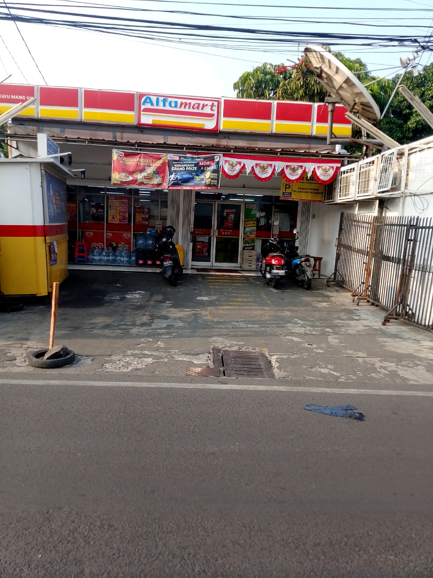 The image depicts the exterior of an Alfamart convenience store, a common sight in East Jakarta, Indonesia. The store features its signature red and yellow signage prominently displayed on the facade. In front of the store, several items are visible: two motorcycles are parked, suggesting customer activity. One motorcycle is dark-colored, and the other is red. A small red stool is also present near the entrance.
The ground in front of the store is paved with concrete and appears slightly wet in some areas, with a visible drain cover. To the left of the store, a yellow and white structure, possibly a kiosk or an extended part of the building, is present. Further to the left, a black tire is placed on the ground, acting as a marker or perhaps a makeshift barrier.
Several banners are hung above the store entrance, advertising promotions and products, including one featuring frozen food and another mentioning a grand prize. A satellite dish is mounted on the roof of the building, and electrical wires are strung overhead, typical of urban infrastructure. A metal fence encloses part of the property to the right of the store entrance. In the foreground, the asphalt road is visible, with a pair of blue jeans lying on it. The environment suggests a typical commercial area in a residential or mixed-use neighborhood of East Jakarta.