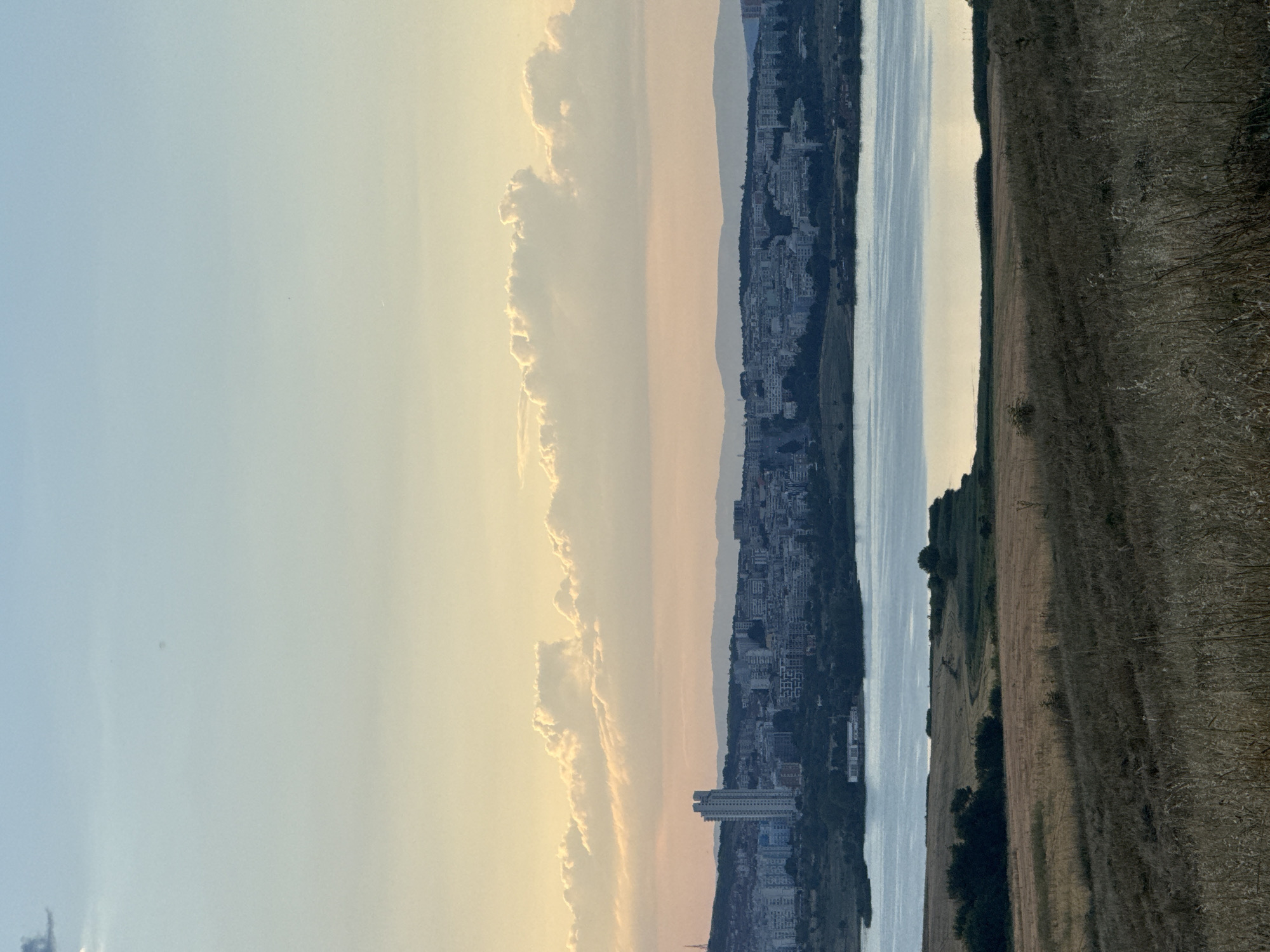 The image depicts a panoramic view of the city of Tvarditsa, Bulgaria, from a slightly elevated vantage point overlooking a body of water and rolling hills. The sky is a gradient of soft blues and pale yellows, with scattered, fluffy clouds illuminated by what appears to be sunset or sunrise light. In the middle ground, a city skyline is visible, characterized by a dense collection of buildings, some of which are high-rise structures. A prominent, slender, light-colored skyscraper stands out among the other buildings on the left side of the city. The city is situated on a hillside that slopes down towards a calm body of water, likely a lake or reservoir, which occupies the mid-lower portion of the image and reflects the soft sky colors. The foreground consists of dry, golden-brown grasses and vegetation, suggesting an arid or late-summer landscape. The overall scene is serene and captures the transition from natural landscape to urban development, with the natural elements dominating the foreground and the city integrated into the distant topography.