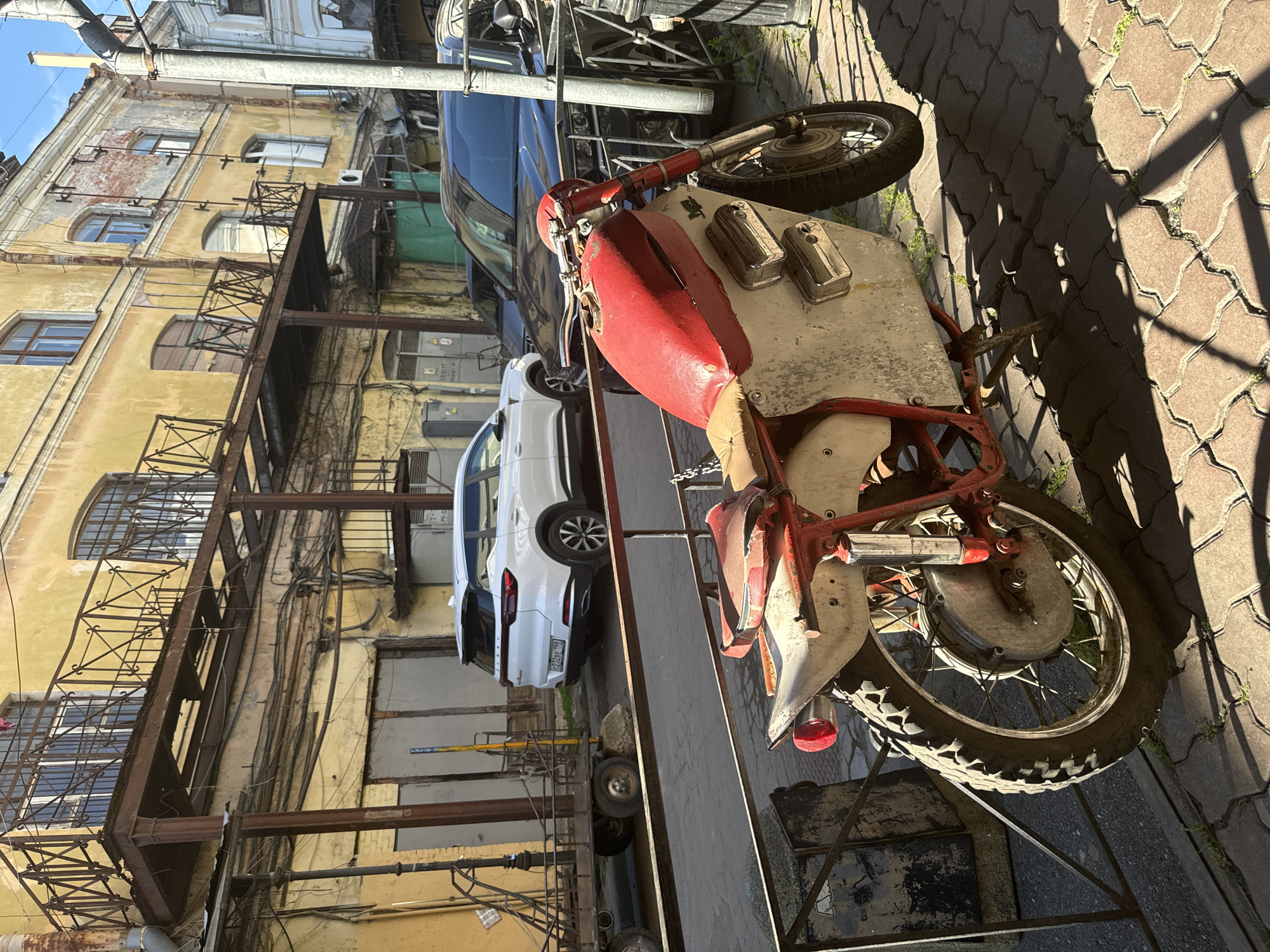 A vintage red and white motorcycle is parked on a cobblestone path in a narrow European-style alleyway. The motorcycle has a rugged, off-road appearance with knobby tires and a high exhaust pipe. It is positioned in the foreground, with its front wheel facing away from the viewer. Behind the motorcycle, a white SUV is parked, with its rear door open, revealing a glimpse of its interior. Further back, a dark blue car is visible. The alley is lined with old, weathered buildings featuring yellow facades, iron balconies, and arched windows. Various pipes and electrical wires run along the exterior walls. The scene is bathed in bright sunlight, casting sharp shadows on the cobblestones, suggesting a sunny day. The overall impression is one of a historic urban environment with a touch of retro charm.