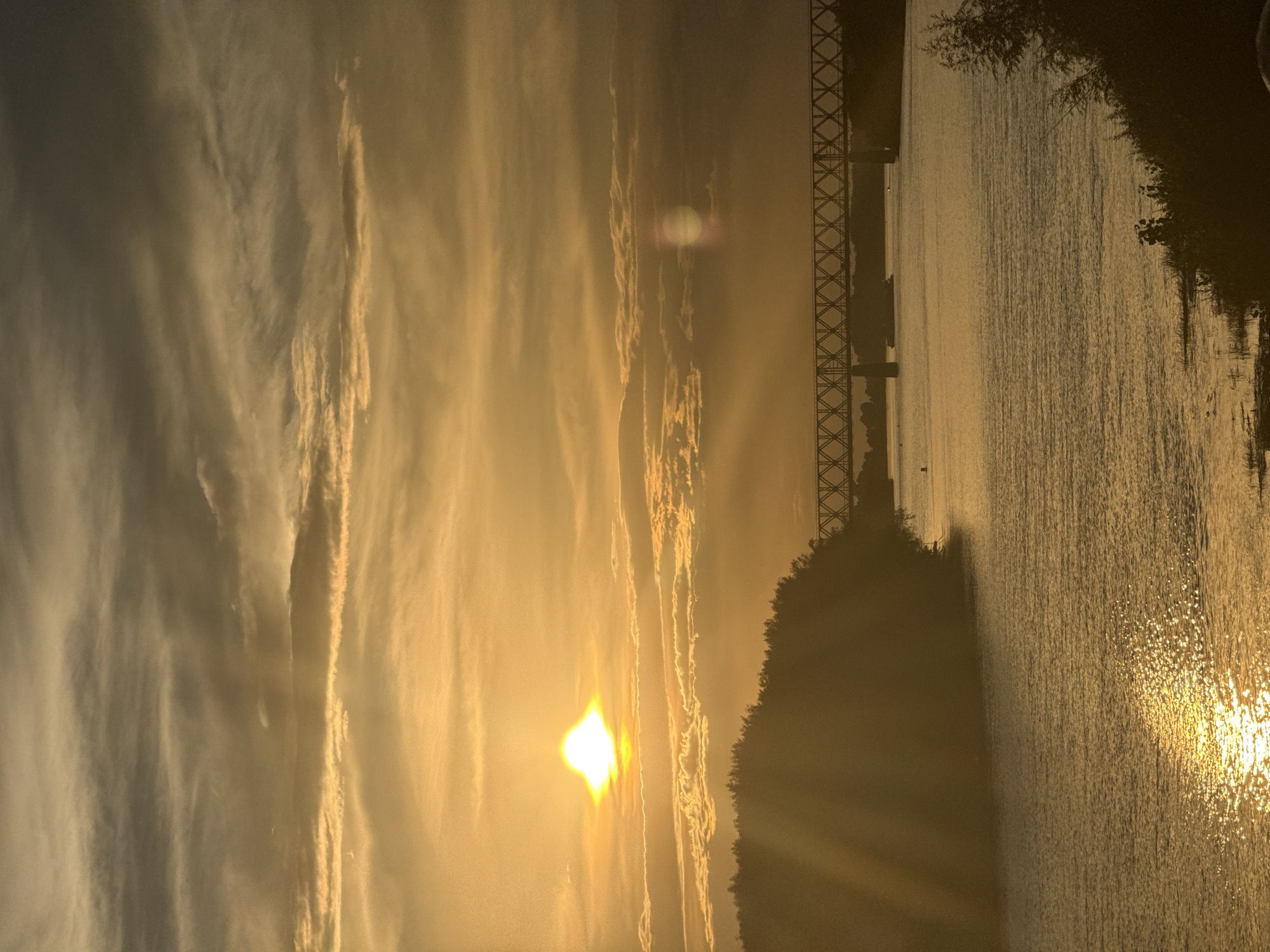 This is a vertical, golden-hour photograph of a wide body of water, likely a river, in Pavlodar, Kazakhstan. The sun is low in the sky, casting a warm, hazy glow over the scene and reflecting brilliantly off the rippling water's surface. Wispy, elongated clouds stretch across the sky, catching the sunlight and adding texture to the atmospheric composition.
On the left side of the image, a silhouette of a tree-lined shore is visible, with a distinct, lattice-work bridge structure extending across the water in the distance. The bridge's metallic framework is clearly defined against the bright sky. On the right, the water dominates the frame, its surface sparkling with sunlight. The overall mood is serene and peaceful, evoking a sense of calm tranquility. The time of day is clearly sunset or sunrise due to the low sun angle and golden light.