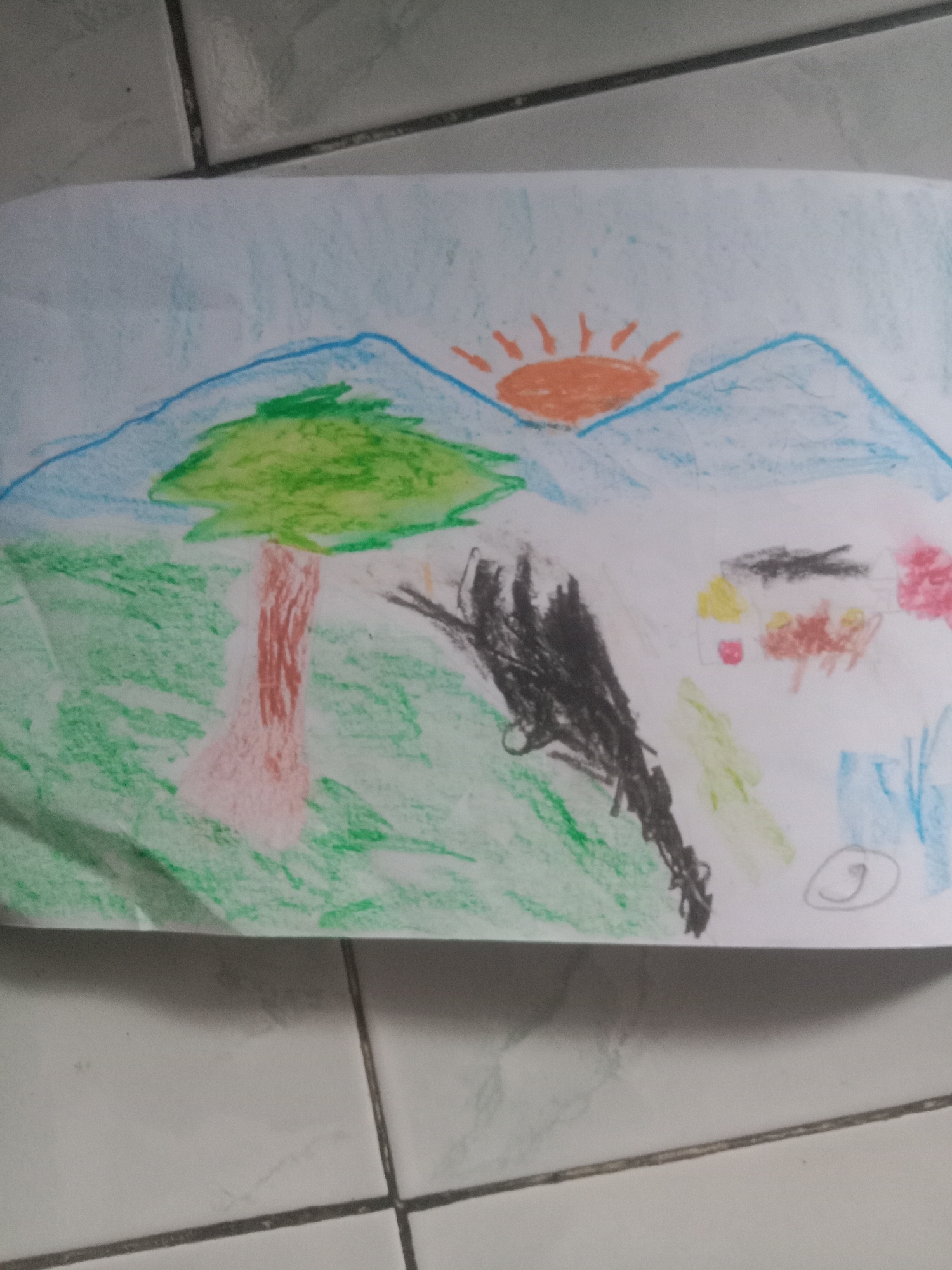 The image displays a crayon drawing on white paper, depicting a landscape. In the background, blue mountains are visible under a light blue sky. An orange sun with radiating lines is situated between the mountains. A large green tree with a brown and pink trunk stands in the middle ground, with green grass filling the lower portion of the drawing. To the right, there are several vaguely human-like figures with yellow, brown, and red coloring, and a pink shape that could be a flower. A black, scribbled object is also present in the middle ground. A circled number "9" is drawn in the lower right corner of the paper. The drawing is placed on a tiled floor. There is no information within the image to suggest a specific location in Kedungsari, Indonesia.