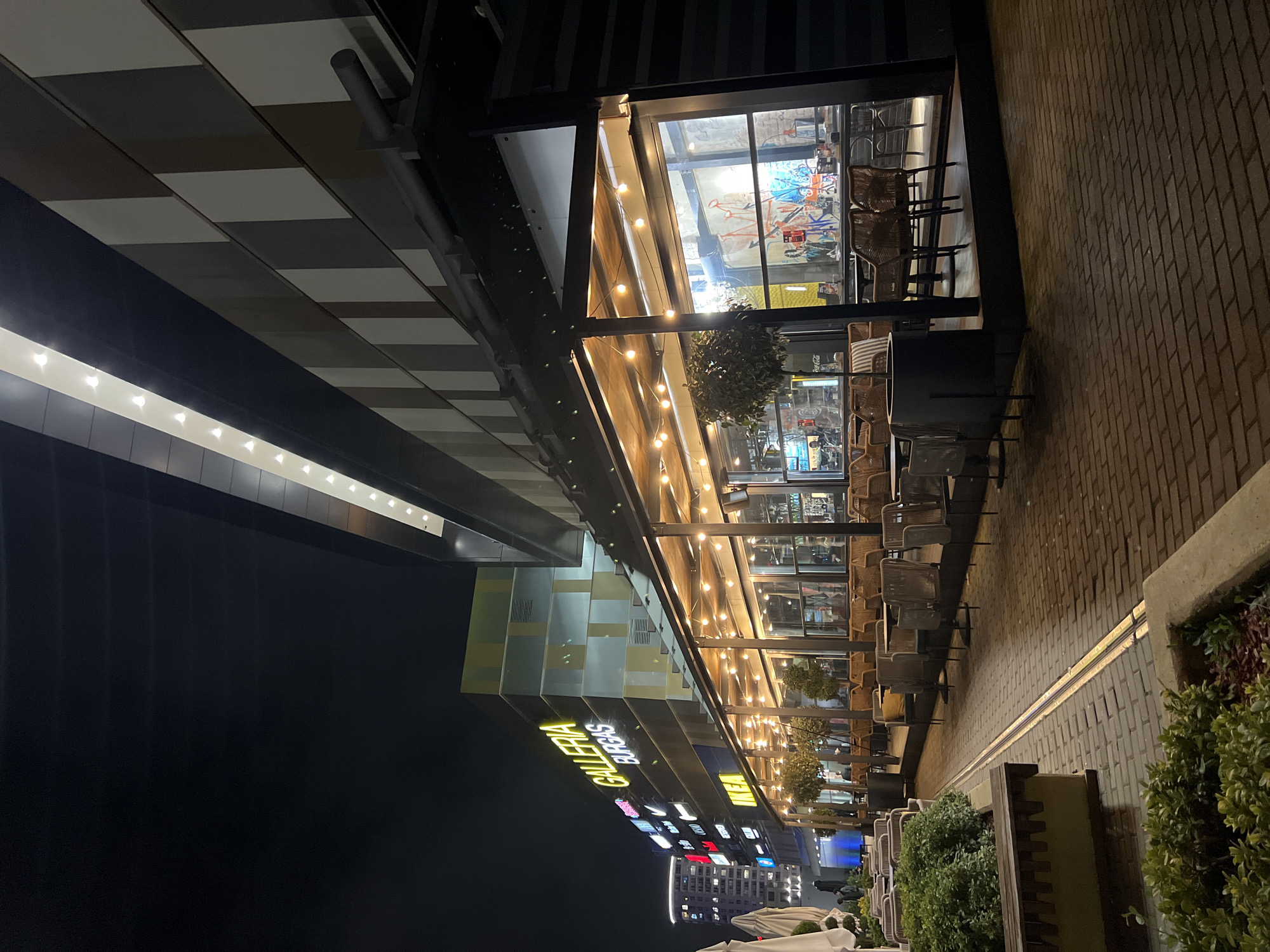 The image shows an exterior shot at night of the "GALLERIA BURGAS" mall in Burgas, Bulgaria. The mall is visible on the left side, with its illuminated signage. A covered outdoor seating area with lights is set up outside the mall on a brick paved walkway. Tables and chairs are arranged in a row adjacent to the mall's glass facade. Green bushes are on the right side, bordering the walkway. In the background, other buildings are visible. The sky is dark, indicating it is nighttime.