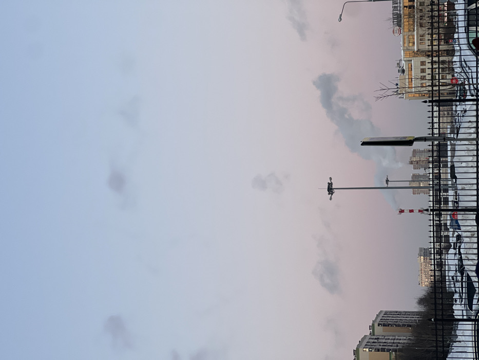 This image depicts a winter urban landscape in Putilkovo, Russia, likely during dusk or dawn, characterized by a pastel sky and widespread snow cover.
The sky transitions from a light blue at the zenith to a soft pink and purple gradient near the horizon. Scattered faint, dark-grey clouds are visible, along with multiple plumes of white-grey smoke originating from sources closer to the horizon.
In the foreground, a black metal fence with vertical bars and pointed finials spans the width of the frame, partially obscured by accumulated snowdrifts. Behind this fence, a snow-covered parking lot contains numerous vehicles, many partially hidden by snow or the fence. A small red "STOP" sign on a white pole is visible within the parking lot area.
From left to right in the midground and background:
On the far left, several multi-story modern residential buildings with light-colored facades are visible. Bare deciduous trees stand between the fence and these structures.
Centrally, a tall metal high-mast light pole with multiple downward-angled luminaires stands prominently.
Further to the right, a distinctive red and white striped industrial chimney emits a vertical column of white smoke. Behind this chimney, additional multi-story buildings, consistent with industrial or commercial facilities, are visible, some also emitting smoke. A large, prominent plume of white smoke rises from this cluster of buildings, drifting horizontally to the right across the sky.
A dark, rectangular vertical sign, possibly a billboard or informational panel, partially obstructs the view in the midground.
On the far right, a standard street light pole with a single luminaire is visible. Adjacent to it stands a multi-story commercial building featuring numerous windows, many appearing illuminated, and rooftop equipment. Signage with Cyrillic text is discernable on its facade.
No individuals are visible in the scene. The presence of snow, smoke plumes, and artificial lighting suggests cold weather conditions and ongoing urban activity.