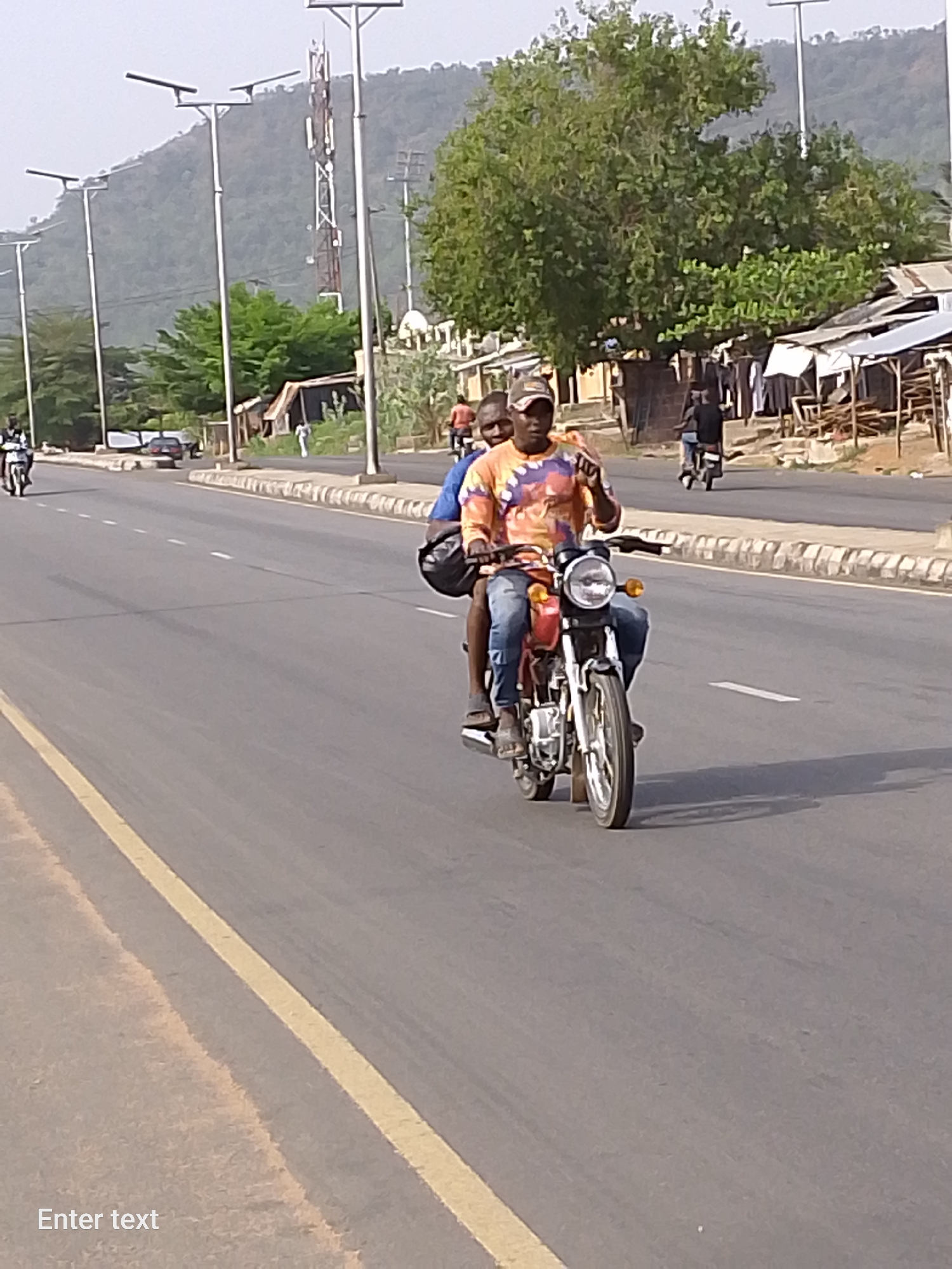 The image depicts a street scene in Adankolo, Nigeria. Two individuals are riding a red motorcycle on a paved road. The driver is wearing a colorful shirt, jeans, and a baseball cap, while the passenger behind is wearing a blue shirt. The driver has his right hand raised. Several other motorcycles and a car can be seen in the background, along with roadside structures and trees. Overhead are streetlights and a telecommunications tower. A mountain is visible in the distance.