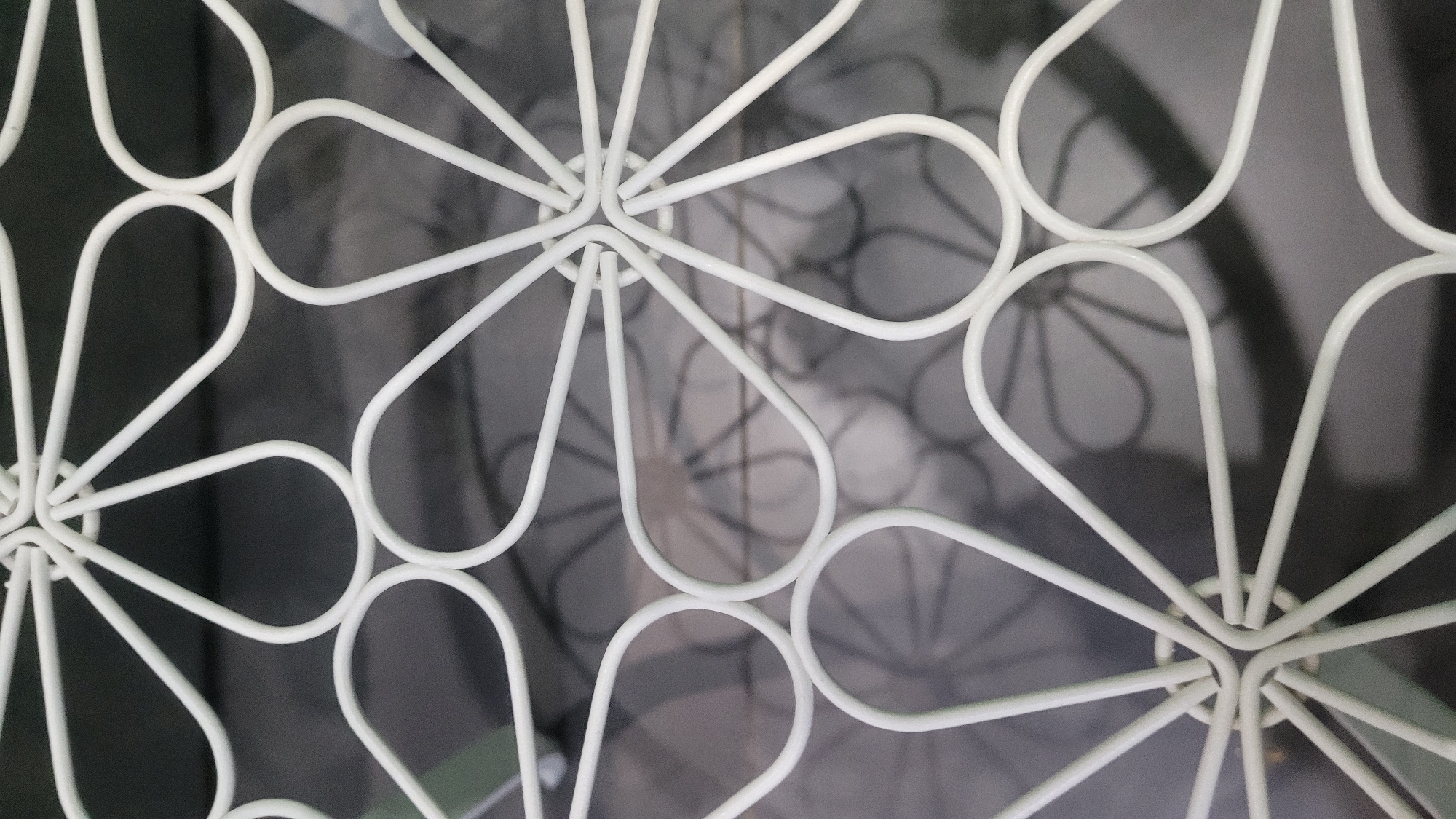 A close-up, overhead view shows a white, wire-frame structure with a floral motif. The structure is made of curved wires that form petal-like shapes radiating from a central point. The background is a dark, textured surface, possibly concrete or asphalt, with faint, shadowy outlines of similar floral patterns. The lighting creates subtle shadows of the wireframe on the background. The scene appears to be outdoors in an urban setting, though the specific location within the city of Unknown, Unknown is not discernible.