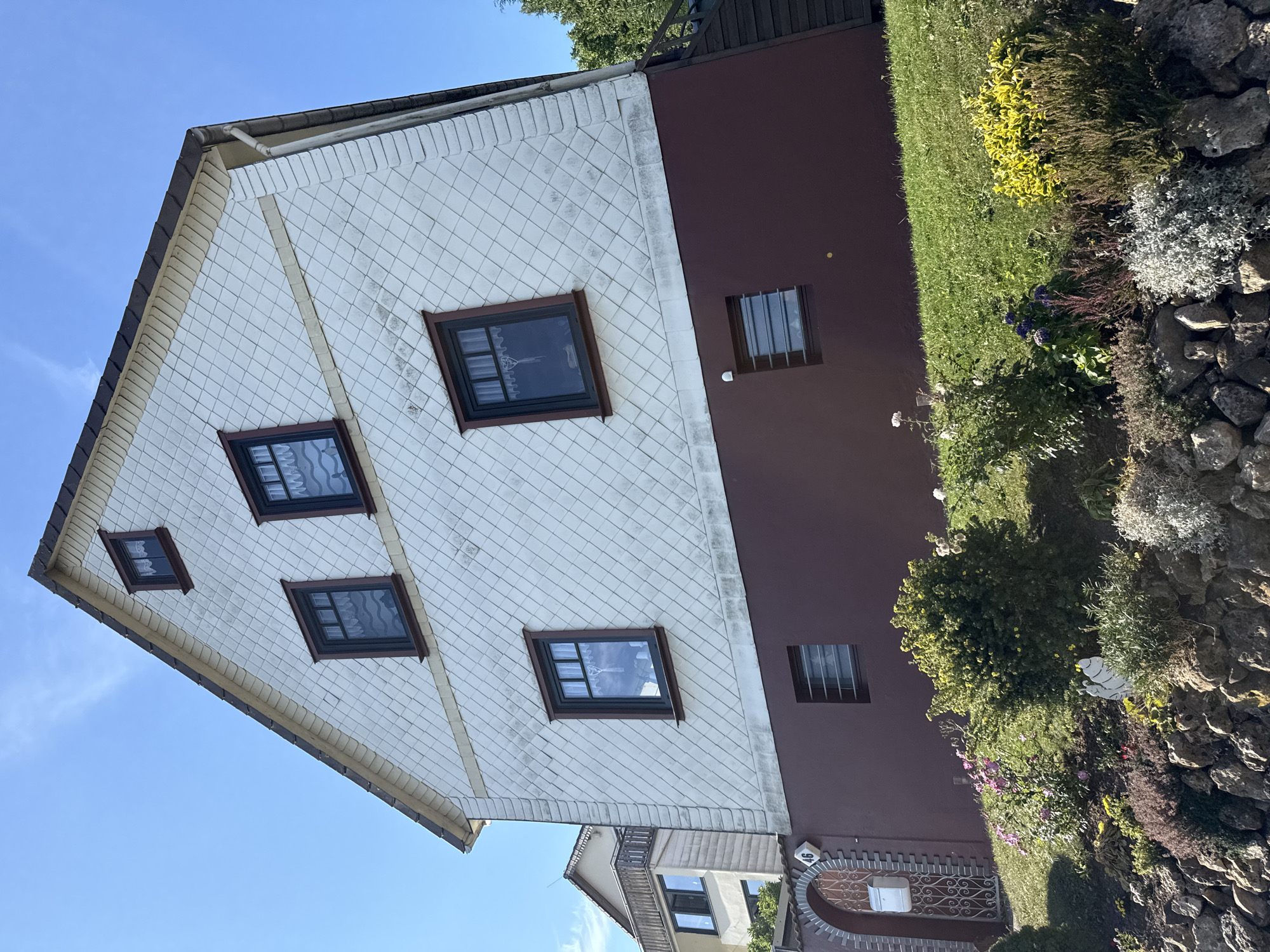 The image shows a multi-story house with a white, diamond-patterned facade on the upper floors and a dark red base. Several dark-framed windows with white lace curtains are visible on the upper floors, along with a small window with bars on the lower level. A pitched roof with dark shingles is visible at the top, and the house is situated on a grassy slope. In the foreground, a garden area with various plants, flowers, and rocks is present. To the left of the house, an arched entryway with an ornate metal gate, a mailbox, and a house number "46" is visible. Another building, possibly an apartment complex, is partially visible to the left of the main house. The sky is clear and blue. The house is located at Ravensberg 46 in Bad Sachsa, Germany. There are no people or animals visible in the image.