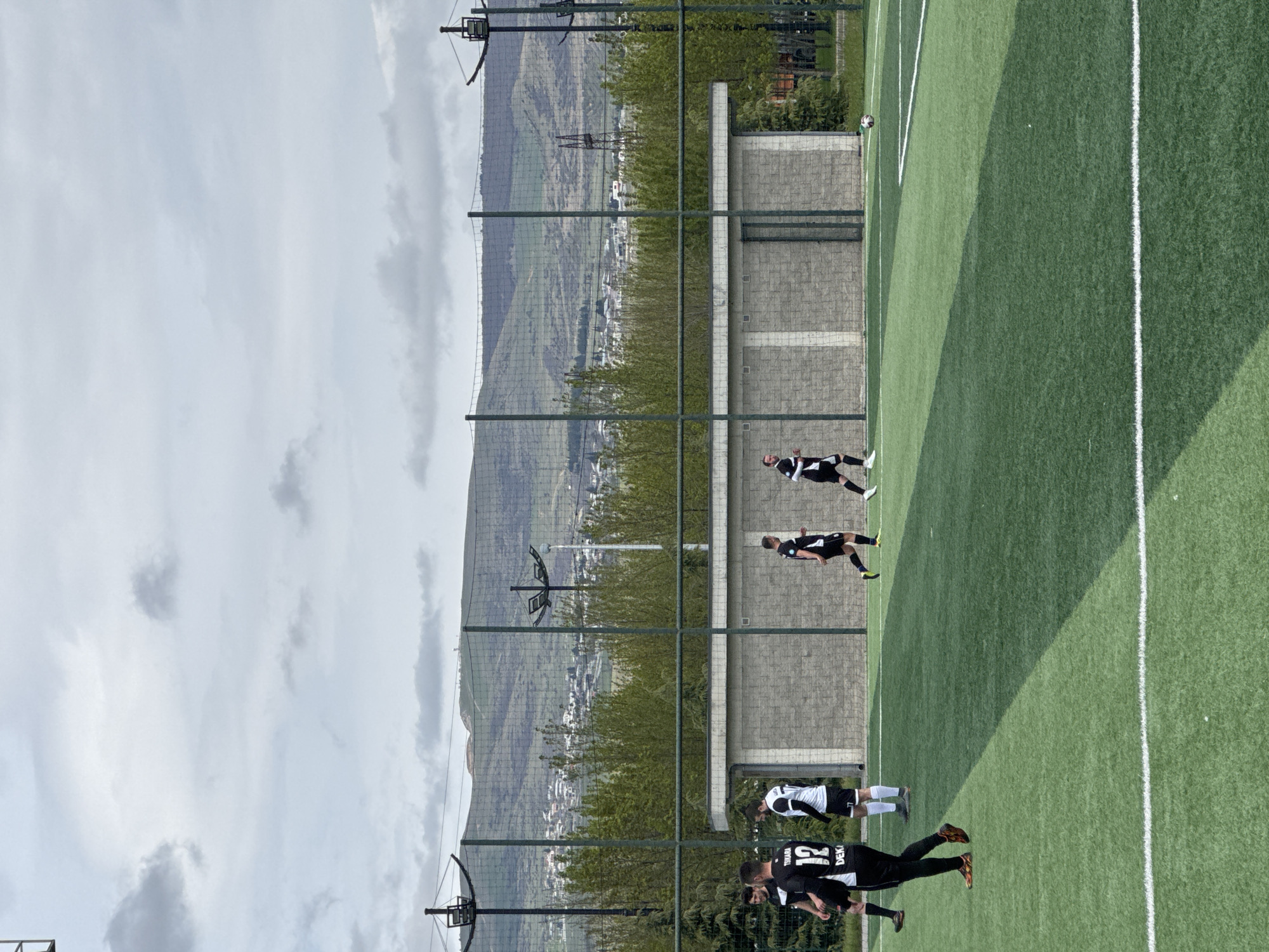 The image shows a soccer field bordered by a high fence and a concrete wall. Several soccer players are visible on the field, wearing black and white jerseys and dark shorts. A soccer ball is in the air near the edge of the field. Beyond the fence, a hilly landscape is visible, with buildings nestled among the trees. The sky is cloudy. It is likely located in Tbilisi, Georgia, which matches the description.