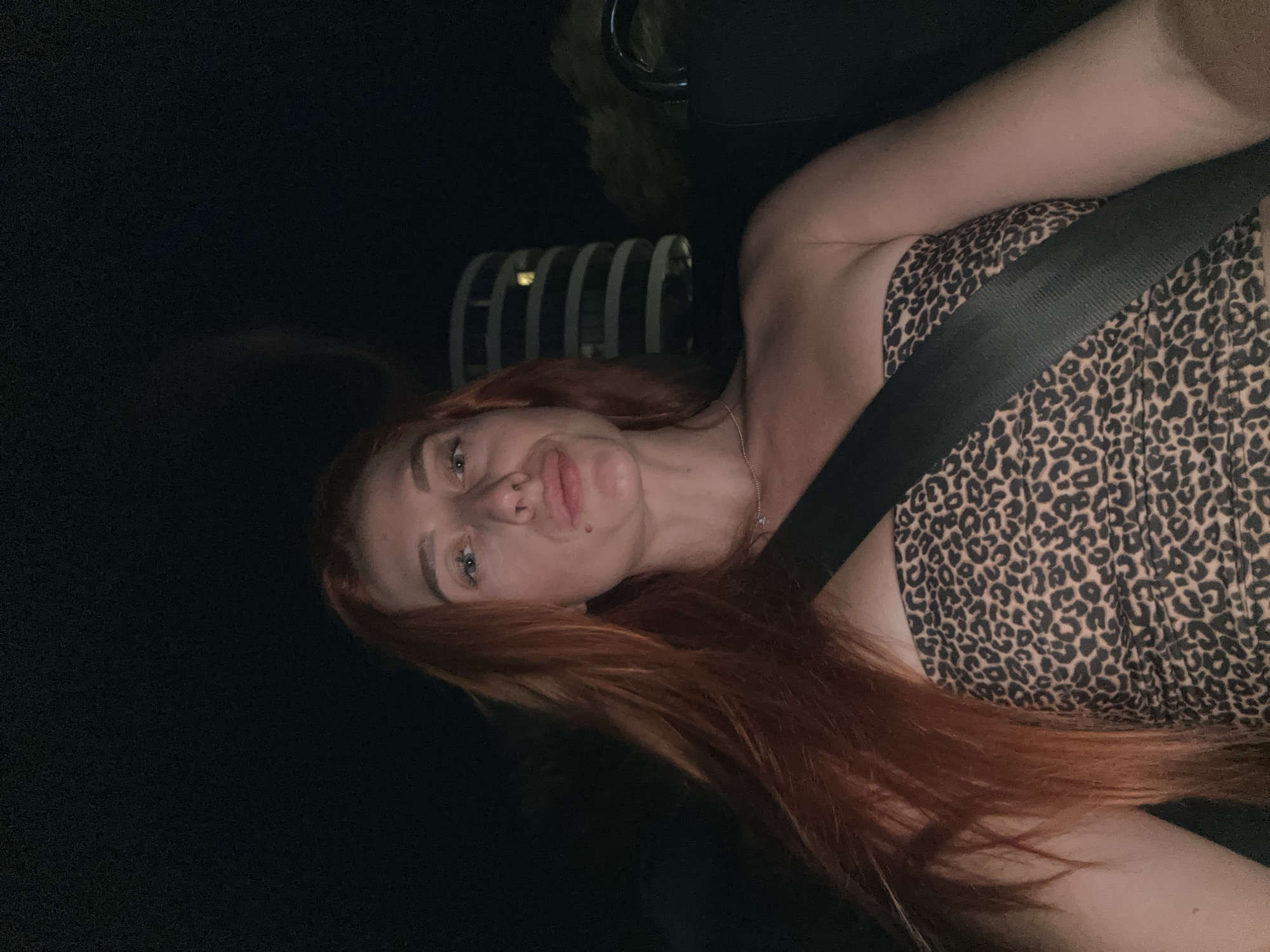 The image shows a young woman with long, red hair sitting in a vehicle at night. She is wearing a strapless, leopard-print top and a black seatbelt. Her head is tilted slightly, and her lips are puckered. In the background, a cylindrical building with illuminated windows is visible. The woman appears to be in the passenger seat, with the seatbelt fastened across her chest. The scene suggests a nighttime car ride through a city, with the building in the background potentially being a landmark in Budva, Montenegro.