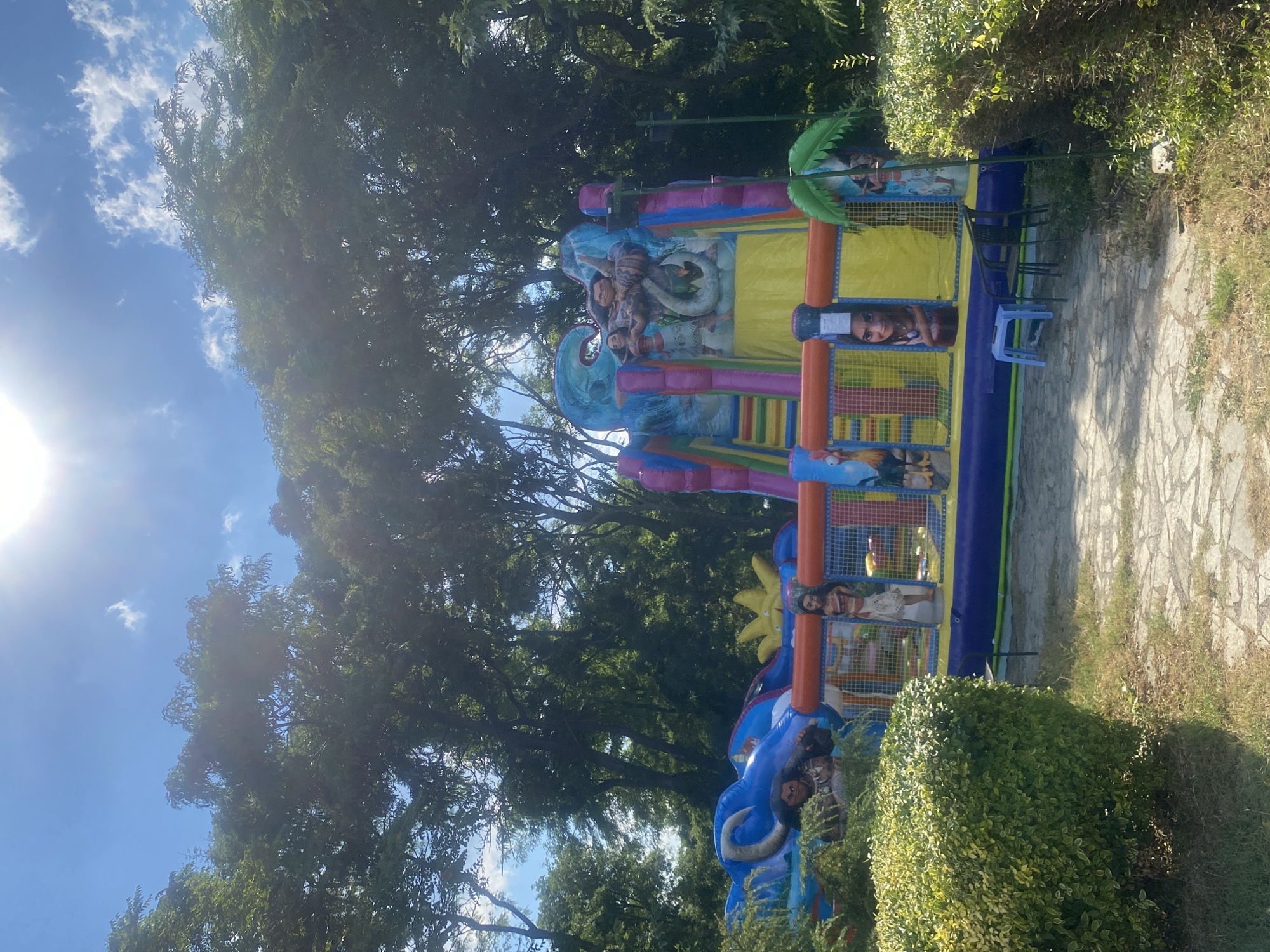 In an outdoor park in the city of Burgas, Bulgaria, a vibrant inflatable playground is set up on a paved stone area. The playground features characters from the Disney movie "Moana," including Moana, Maui, and Heihei the rooster. The inflatable structure is colorful, with blues, yellows, pinks, and purples dominating its design. It includes slides and climbing areas. Lush green trees form a dense backdrop behind the playground, and the sky above is blue with scattered white clouds and bright sunlight. In the foreground, manicured green bushes partially obscure the view of the playground. A light blue plastic stool and two dark chairs are placed near the playground entrance.