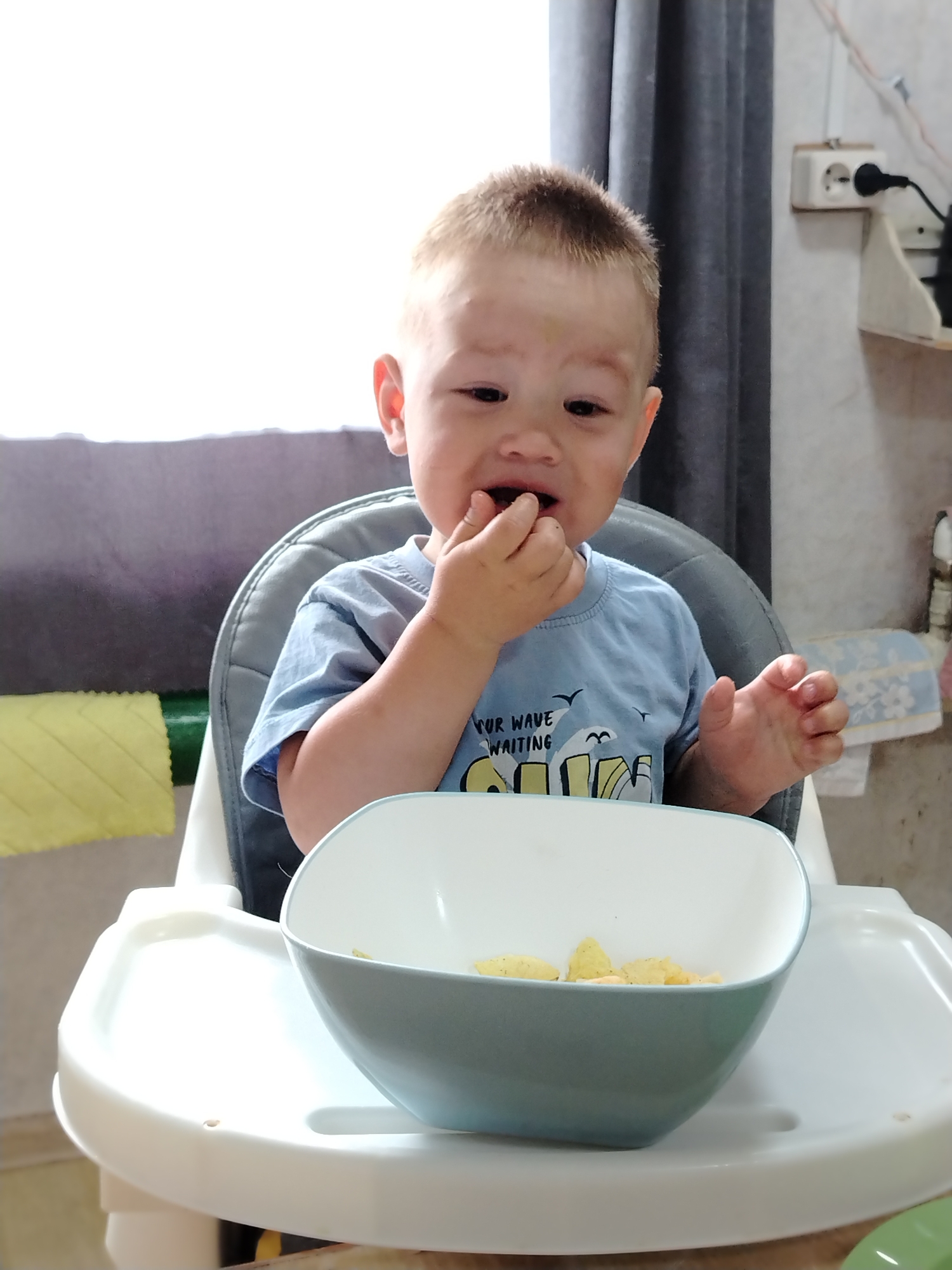 A young child is sitting in a highchair, eating from a blue bowl. The child has short, light brown hair and is wearing a light blue t-shirt with a graphic print. The highchair is predominantly white with a gray backrest. The bowl contains what appear to be yellow snack pieces. The background includes a gray curtain, part of a window, and a wall with an electrical outlet and a shelf with items on it. The location is indoors in a home setting, within the city of Unknown, Unknown.