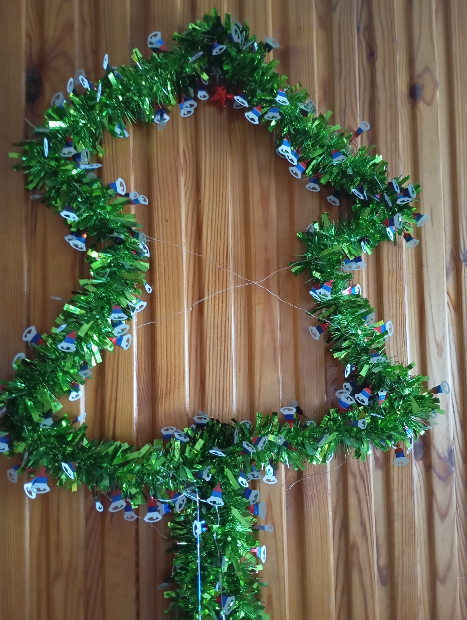 A Christmas tree-shaped garland of green tinsel is hanging on a light brown wooden wall. The garland is adorned with small, white, bell-shaped ornaments, some of which have red and blue accents, resembling the Russian flag. The garland appears to be made of shiny, metallic material and is illuminated by a string of small lights, also incorporated into the garland. There are thin, white threads, likely part of the garland's structure or meant for hanging, visible against the wooden background. The overall impression is of a festive, seasonal decoration. The image does not contain any people or provide explicit geographic information beyond a general context of being indoors on a wooden wall.
