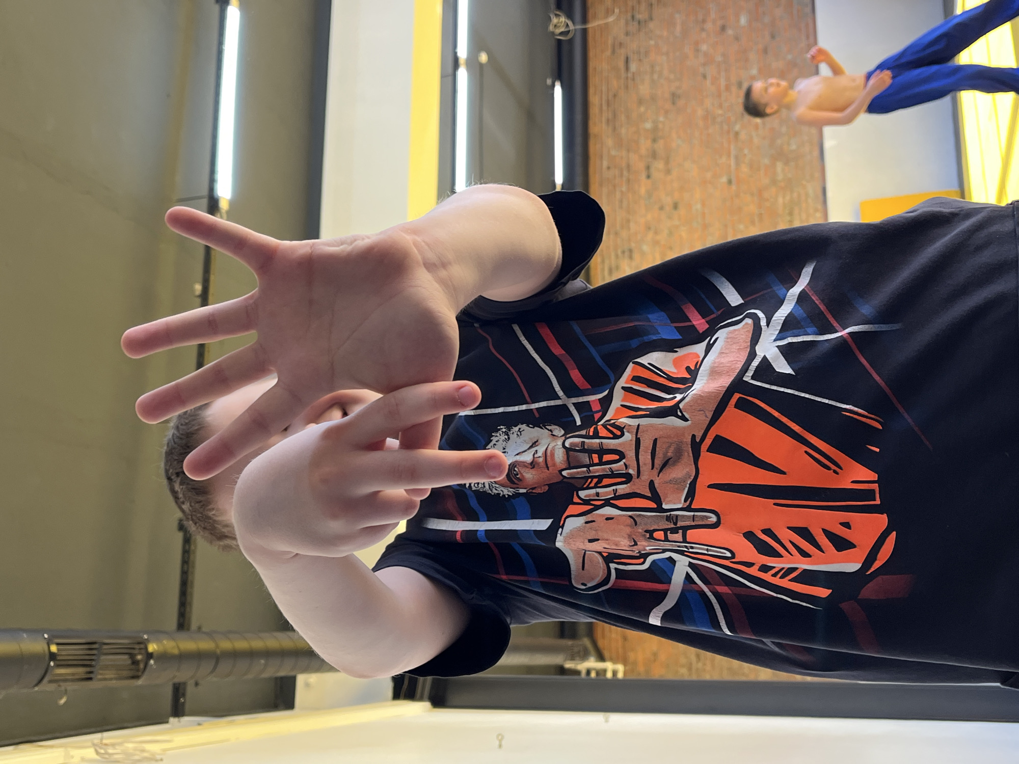 The photo shows a person wearing a black t-shirt with a graphic print and holding their hands up towards the camera. The background includes elements of an indoor setting, such as lighting fixtures and a brick wall. The scene depicts a statue of a person in the background that is in the city of Krasnoyarsk, Russia, at the Geo.Mirov Exhibition Center.