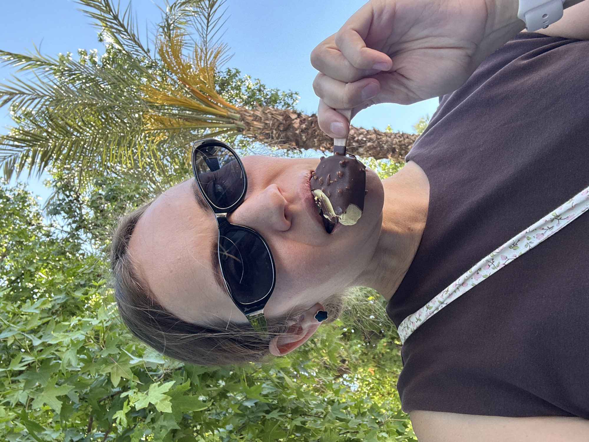 A woman wearing sunglasses and a brown shirt is holding an ice cream bar in front of her face with her right hand. The ice cream is covered in chocolate and nuts with a bite taken out of it. The background features trees, including a palm tree, and a clear blue sky. The woman is standing outdoors in Fethiye, Turkey.