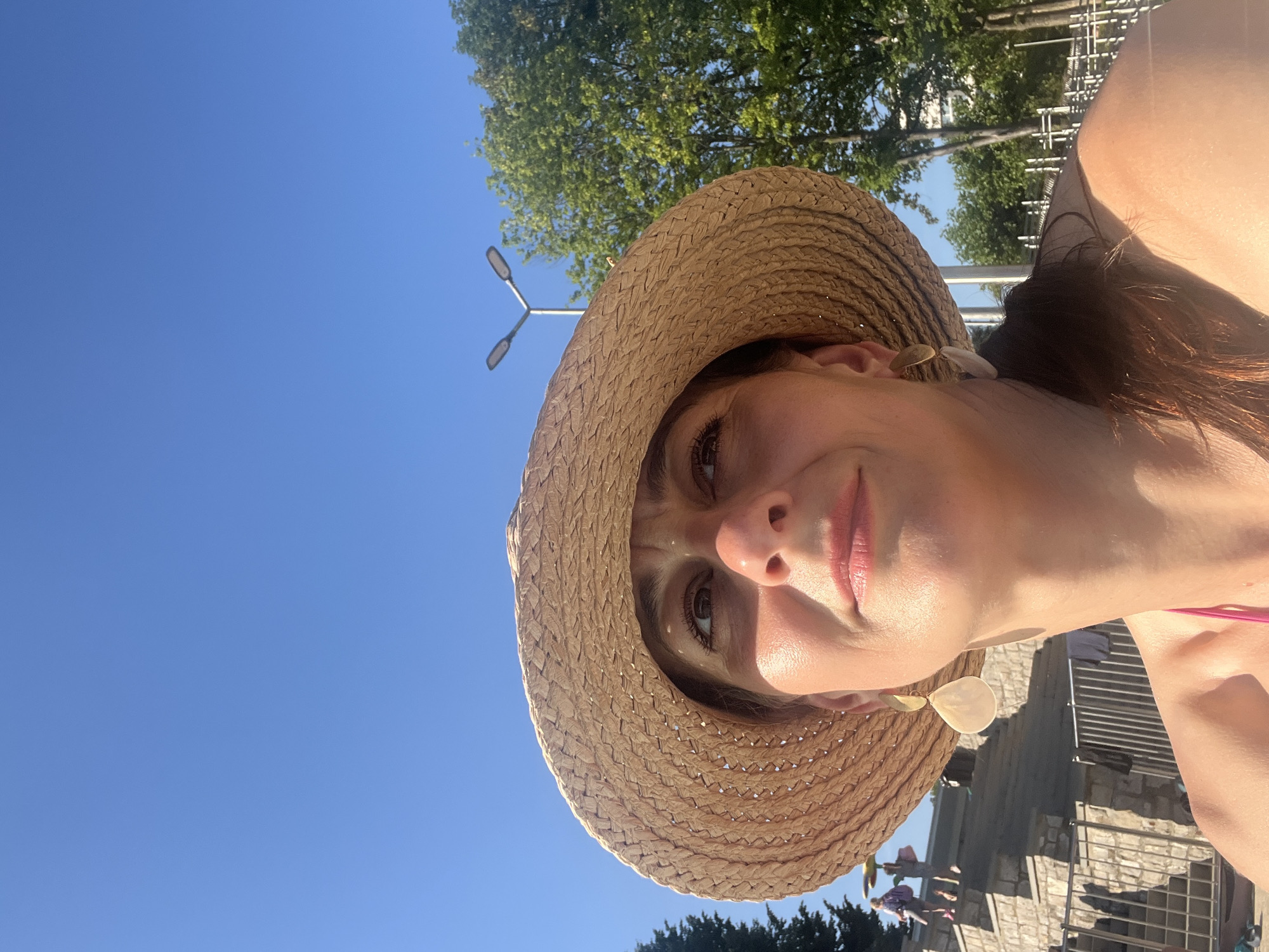 The image is a selfie of a woman wearing a straw hat. She is outdoors with a clear blue sky in the background, and the sun is shining brightly, suggesting it is daytime. Trees and some buildings are visible in the background, giving a hint of a park or a public outdoor space. The woman is looking at the camera with a slight smile. She is wearing a pink strap, possibly from a swimsuit or a top. There is a large, light-colored earring on her right ear. The overall mood is casual and relaxed, suggesting a day of leisure.