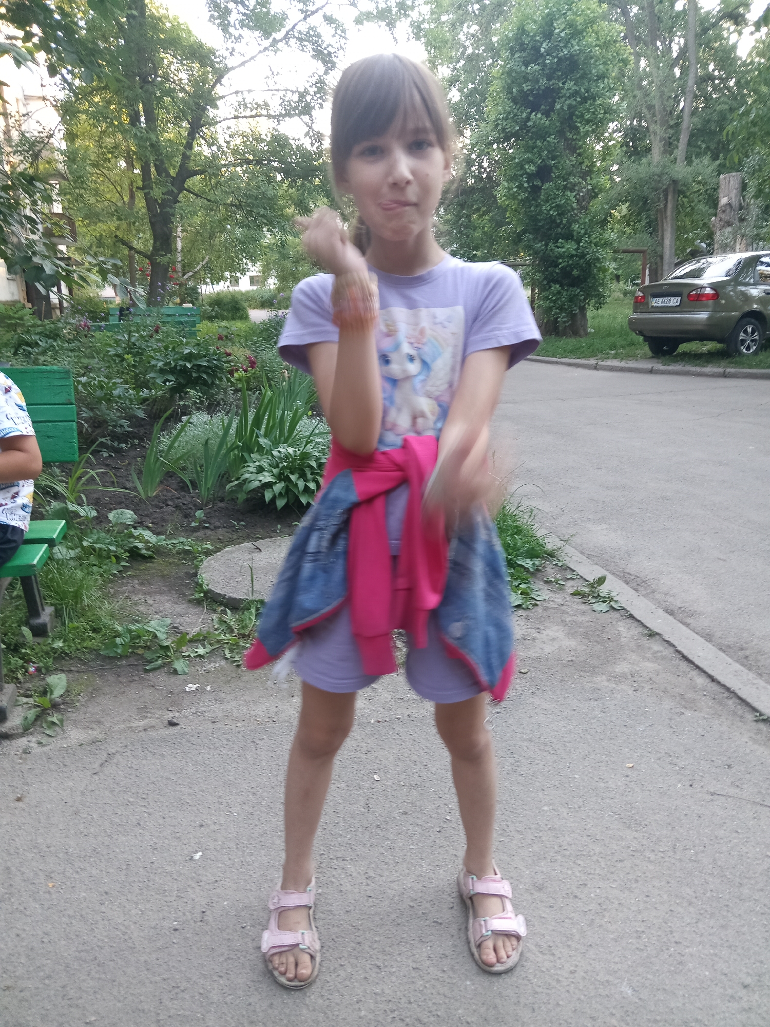 A young girl stands in a residential area. She has brown hair and is wearing a purple t-shirt with a unicorn graphic, a pink shirt tied around her waist, and denim shorts. She is also wearing pink sandals. The girl is facing the camera with her tongue sticking out. In the background, there are trees, bushes, a car, and a building.