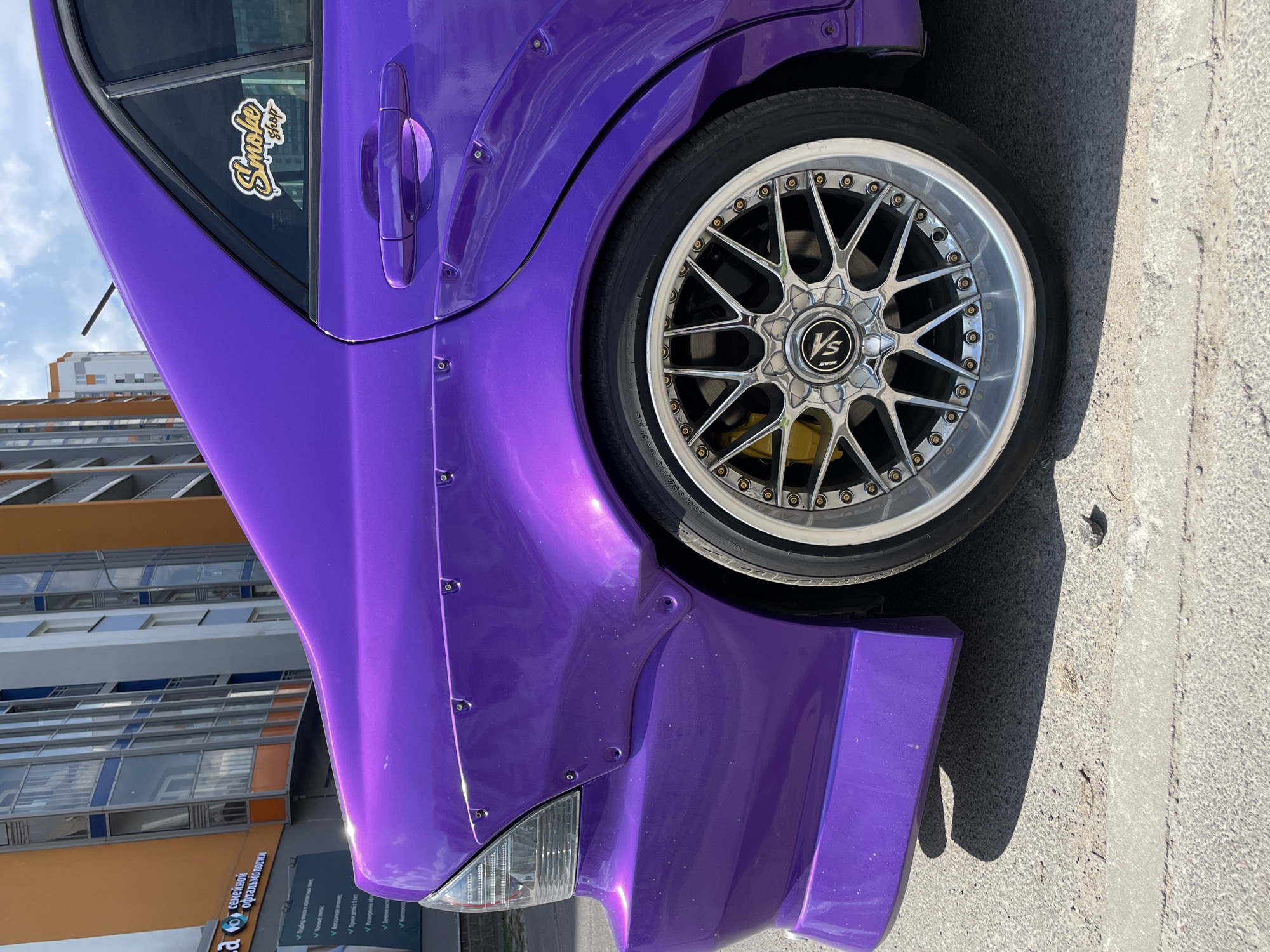 The image depicts the rear-left quarter of a modified purple car parked outdoors under bright daylight in an urban environment. The vehicle is a sedan, likely a Lexus IS or Toyota Altezza, characterized by its metallic purple paint finish. Visible modifications include widebody fender flares attached with numerous small, silver rivets or bolts, extending from the rear bumper line over the wheel arch to the rear door.
The car features multi-spoke aftermarket wheels with a polished lip and dark centers, and yellow brake calipers are visible through the spokes. A purple aftermarket rear bumper extension or skirt is present. The rear-left tail light, partially visible on the far left, has a clear or white housing with horizontal lines. The rear-left door handle is also visible, painted to match the car's body. On the rear-left window, a yellow and black stylized sticker reads "Smoke shop." A slender black antenna rod extends upwards near the rear window.
In the background, two multi-story buildings are visible. The building on the left has an orange facade with numerous large, rectangular windows, some framed in blue. A sign below the windows displays Cyrillic text, which translates to "family ophthalmology," suggesting a Russian-speaking location. To the right and further back, another taller building with a grey or white facade and balconies or windows is present. The sky above is partly cloudy blue. The car is parked on an asphalt surface showing cracks and texture, with a visible white painted line. A shadow from the car is cast on the ground to the right. No people are visible in the scene. The precise location in the city of Unknown, Unknown is not identifiable beyond the linguistic hint provided by the sign.