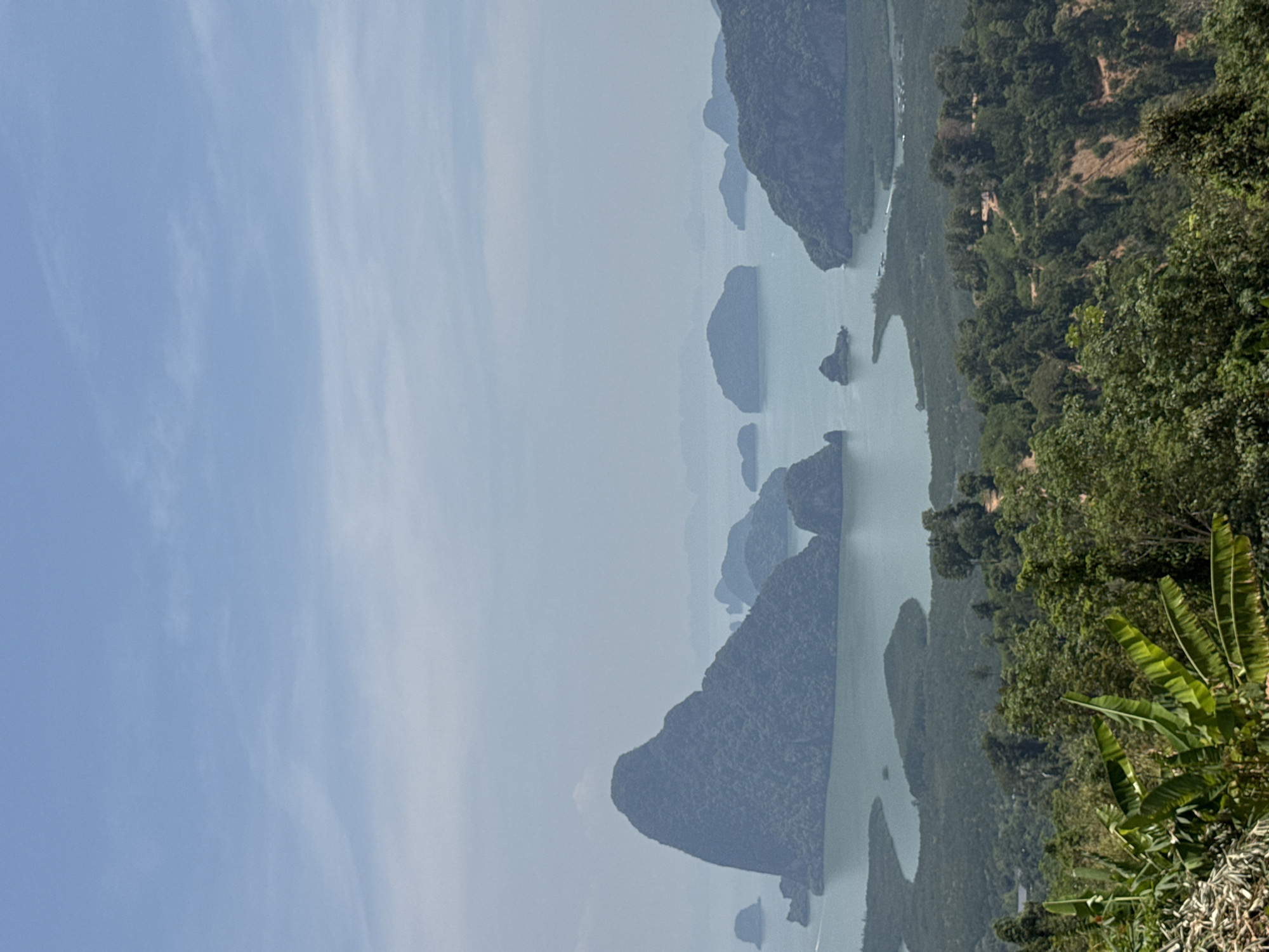 The image shows a landscape in Ban Khlong Khian, Thailand, featuring a bay dotted with limestone karst formations. The water is a light green-grey color. Lush green vegetation covers the edges of the land and some of the karst islands. The sky is light blue with some clouds or haze. The view is from an elevated position, possibly a viewpoint, looking down on the bay. Banana plants are in the foreground.