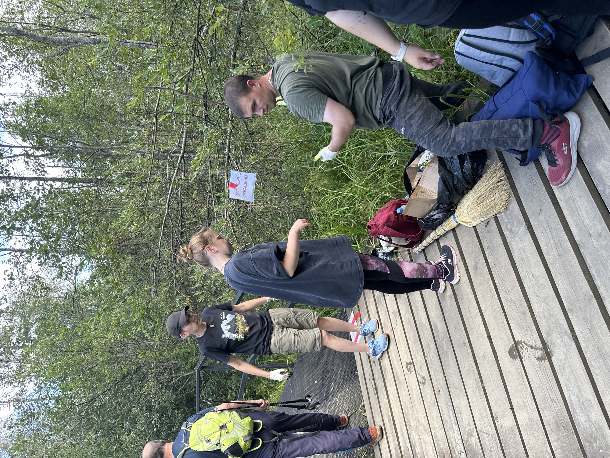 Five individuals are on a wooden boardwalk amidst a dense forest. A man on the left, wearing a bright yellow backpack and grey pants, holds two trekking poles. Next to him, a woman in a black t-shirt and khaki shorts wears a black cap. In the center, a young woman in a dark oversized t-shirt and patterned leggings stands. To the right, a man in a green t-shirt and camouflage pants is holding yellow gloves and a green object, possibly a tool. Another person, partially visible on the far right, wears a black t-shirt and has a white watch. Several garbage bags, a broom, and backpacks are on the boardwalk. A white sign with red lettering is posted on a tree in the background. The environment appears to be a nature trail or park area.