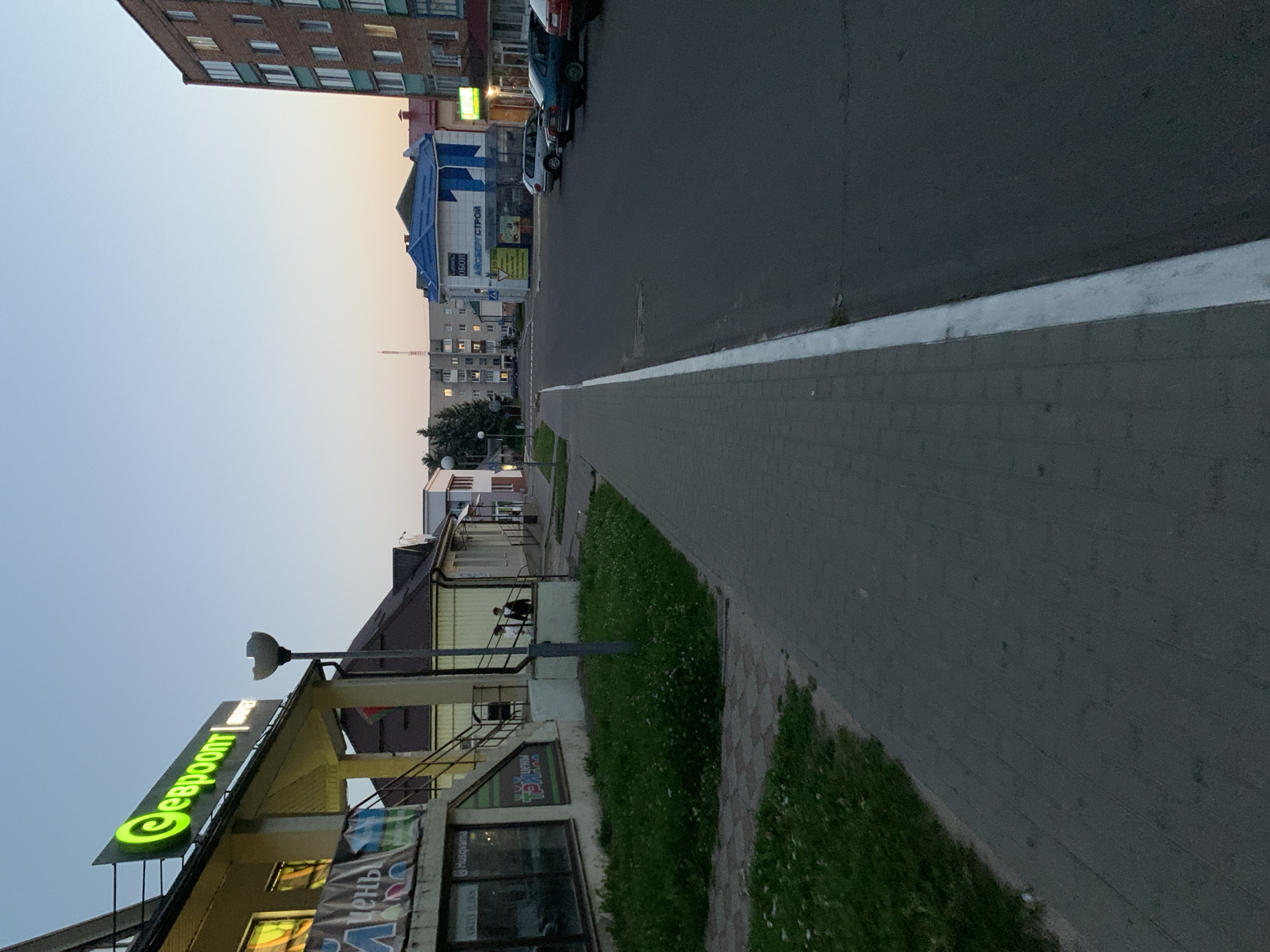 The image depicts a street scene in Kapyĺ, Belarus, likely during the evening or early morning given the soft, diffused light. The foreground features a paved sidewalk constructed of interlocking gray bricks, bordering a strip of well-maintained green grass dotted with small white flowers. To the left of the sidewalk, a low building houses a store with large windows, displaying various advertisements and signage. One prominent sign features a bright green logo with Cyrillic text, "евроопт," which translates to "Evroopt," a popular Belarusian supermarket chain. Adjacent to this is another banner with colorful graphics and Cyrillic text. A dark-colored staircase with a metal railing leads up to the store entrance.
Further down the sidewalk, a streetlamp with a distinctive, broken dome-shaped light fixture stands tall. Two individuals, a man and a woman, are seen walking up the stairs towards the building.
The street itself is paved with asphalt and marked with a white line along the edge of the sidewalk. Several cars are parked along the street. In the middle ground, a building with a bright blue roof is visible, housing a store with Cyrillic signage that appears to indicate building materials or home improvement supplies, with text like "ОБОИ" (wallpaper) and "СТРОЙ" (construction). Further in the background, multi-story apartment buildings and other structures of varying architectural styles line the street, suggesting an urban or semi-urban environment. The sky is a pale, clear gradient from a light blue near the horizon to a deeper blue above, indicating the time of day.