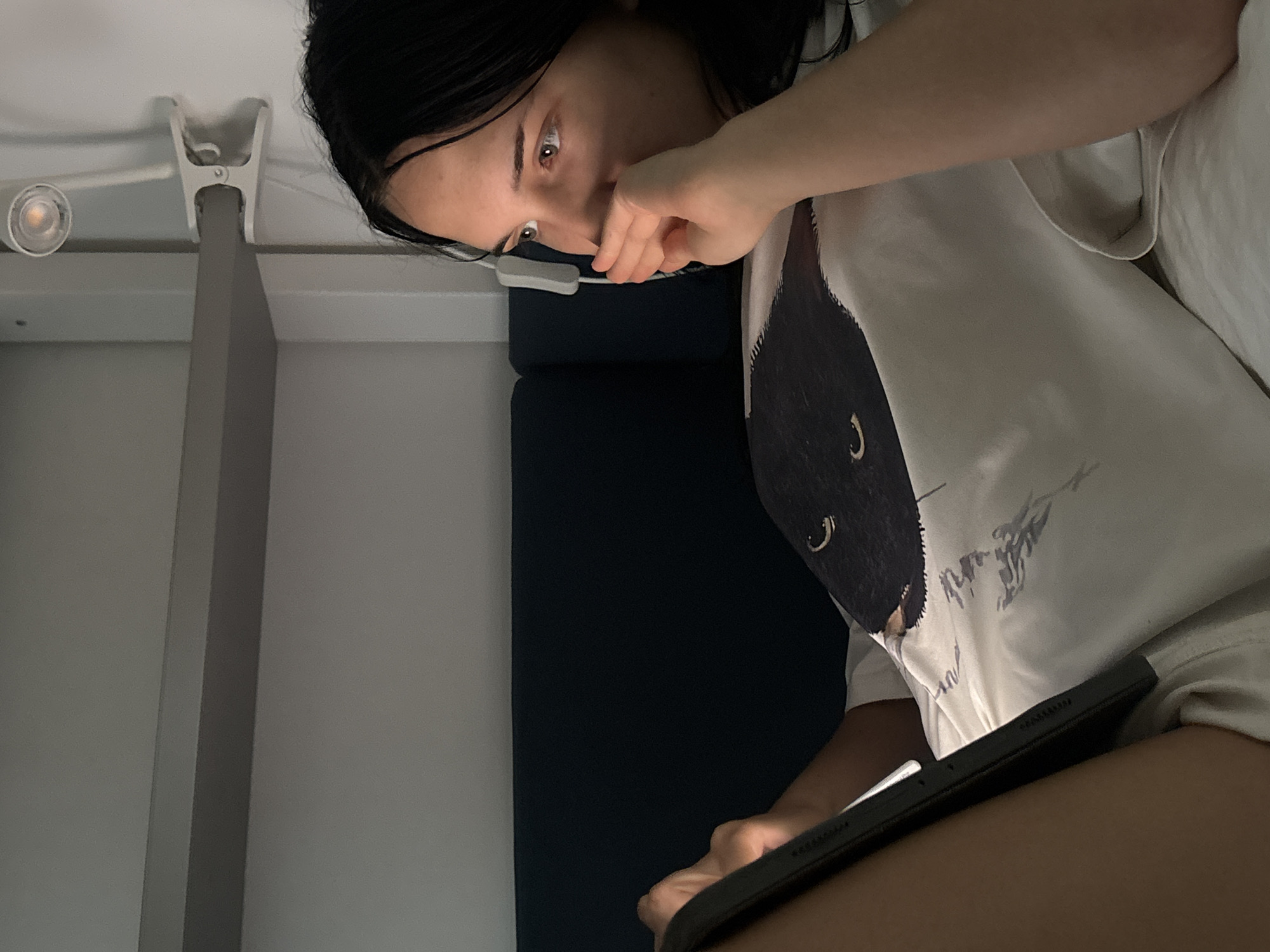 A young woman with dark hair is lying down in bed, looking at a tablet device. She is wearing a white t-shirt with a black cat graphic on it. Her right hand is raised to her mouth, and she appears to be deep in thought or concentration. The room is dimly lit, with a clip-on lamp providing light. The background shows a dark blue headboard and a light grey shelf. The scene suggests a private, indoor setting, possibly a bedroom. There is no information available to associate this image with Cupra Marittima, Italy.