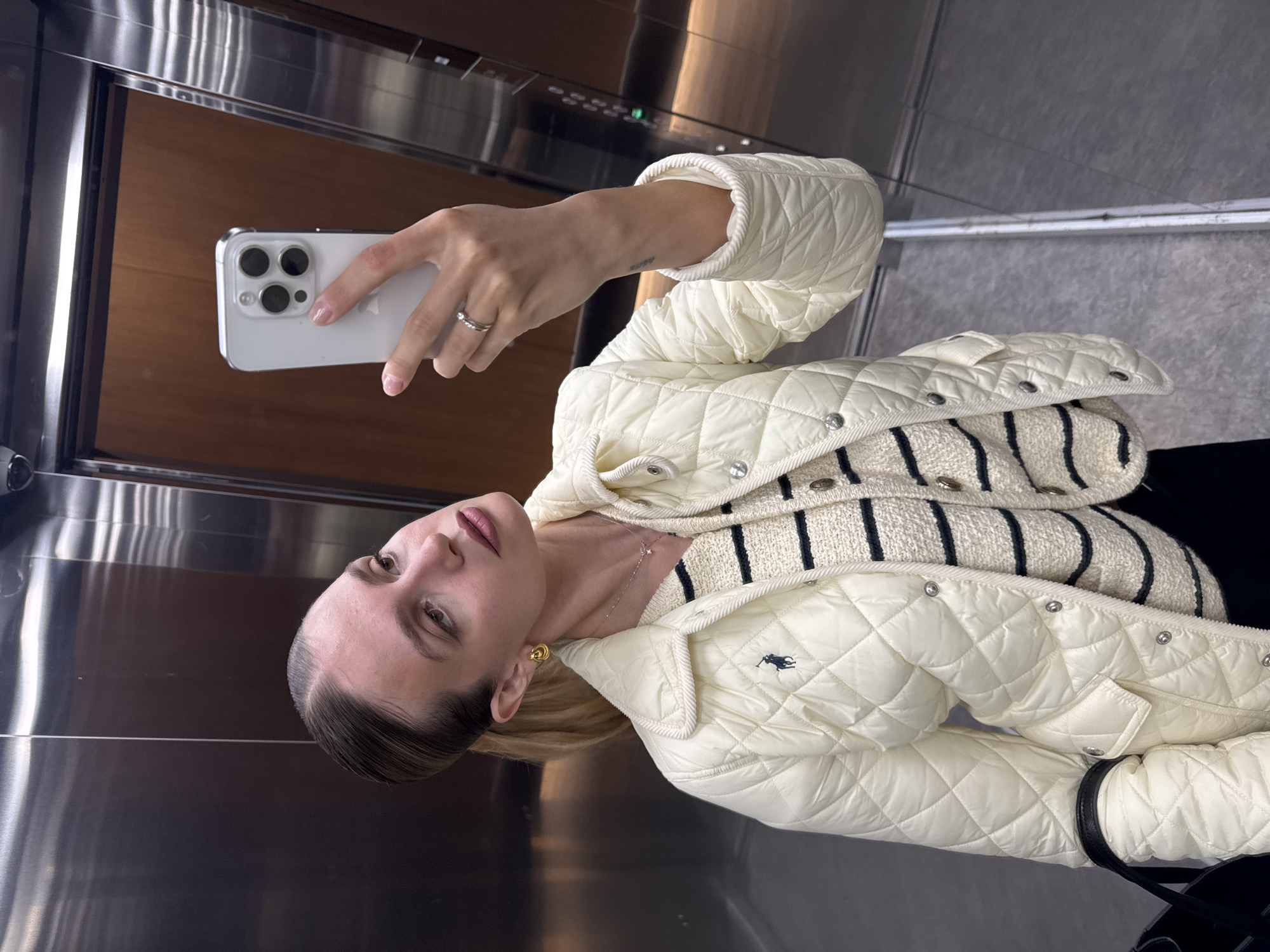 A woman is captured in a mirror selfie, holding a white iPhone 14 Pro with a clear case in her right hand. She has light brown hair pulled back and wears a neutral expression with subtle makeup, including pink lipstick. She is dressed in a cream-colored quilted jacket with a collar and snap buttons, featuring a small dark blue Polo Ralph Lauren horse logo on the left chest. Beneath the jacket, she wears a cream-colored knitted sweater with black horizontal stripes.
Her accessories include a multi-band silver ring on her right ring finger, small gold stud earrings, and a delicate silver necklace with a tiny pendant. A faint "luv" tattoo is visible on her inner right forearm.
The setting is an indoor space with highly reflective surfaces, likely an elevator or a mirrored hallway. The reflections show polished metallic panels and warm wooden textures, suggesting a modern and clean environment. The ceiling is visible as a grey surface. The visible text includes the Polo Ralph Lauren logo on her jacket and the Apple logo on the back of her phone.