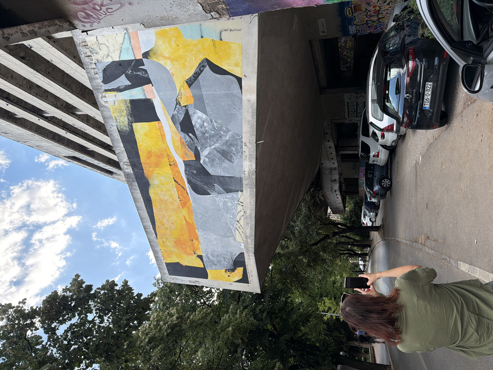 In Mostar, Bosnia and Herzegovina, on a sunny day with scattered clouds, a woman with auburn hair stands on a paved road, facing away from the camera. She is wearing a green shirt and holding a black smartphone in both hands, appearing to take a photograph. In the background, an abstract mural adorns the side of a large concrete building. The mural features geometric shapes and a color palette of yellow, orange, gray, black, and white, with some pastel colors like light blue and pink. The building appears to be an elevated structure, possibly part of a parking garage or an unfinished building, with visible concrete beams and some graffiti on the lower sections. To the left of the woman, a line of trees with dense green foliage creates a canopy over the sidewalk. Further down the road, several cars are parked along the side, including a black Renault Megane with a license plate reading "TD6-0-822." The environment suggests an urban setting with a mix of modern architecture and natural elements.