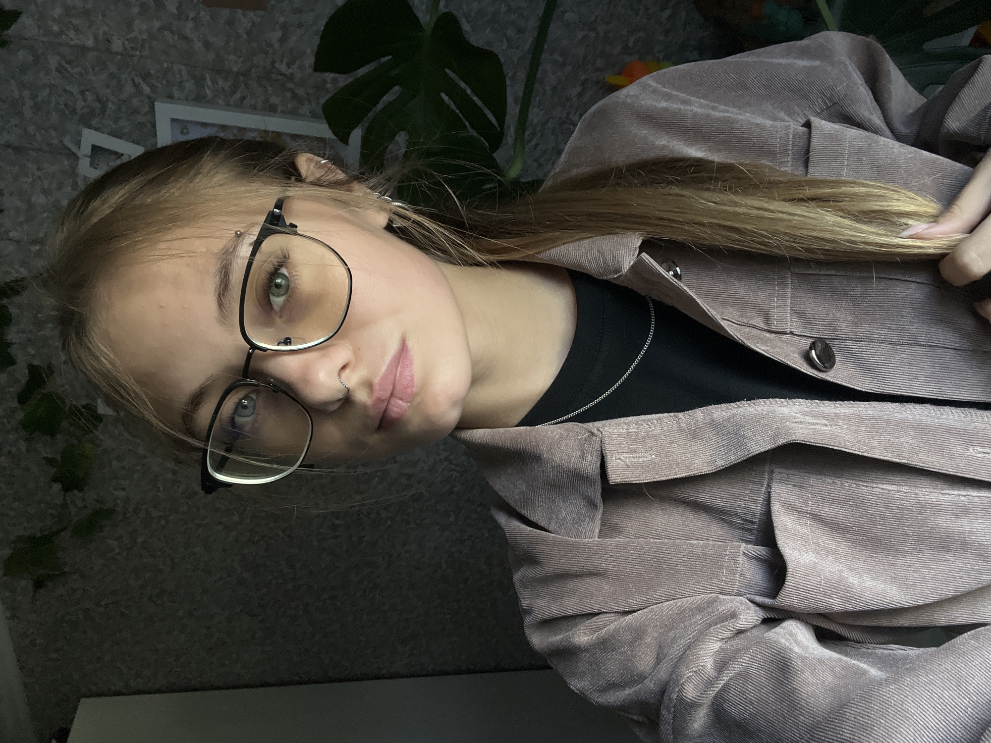 A young woman with blonde hair pulled back into a ponytail is looking at the camera. She is wearing glasses with black frames, a nose ring, and a silver necklace. She is also wearing a brown corduroy jacket over a black shirt. The background is a dark, textured wall with some green plants visible. A large leaf of a Monstera plant is prominent to the right of the woman. The lighting suggests an indoor setting, possibly a home, with a light source coming from the left. The image appears to be a selfie. The location of Фоки, Russia is not discernable from the provided image.